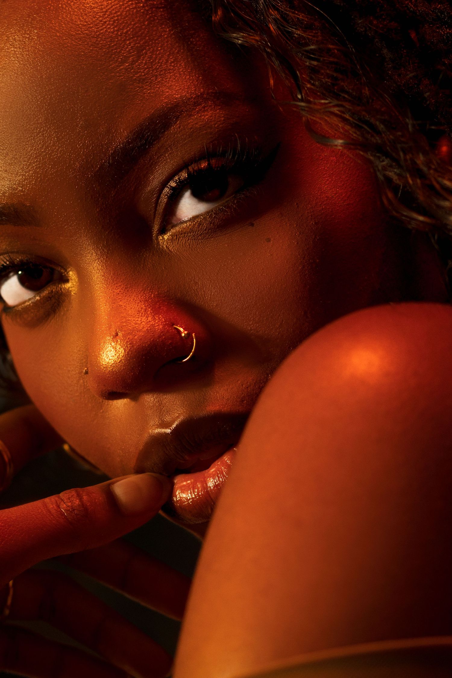 Close-up portrait of a Black woman with nose ring and gold eye makeup, finger near lips in warm light.