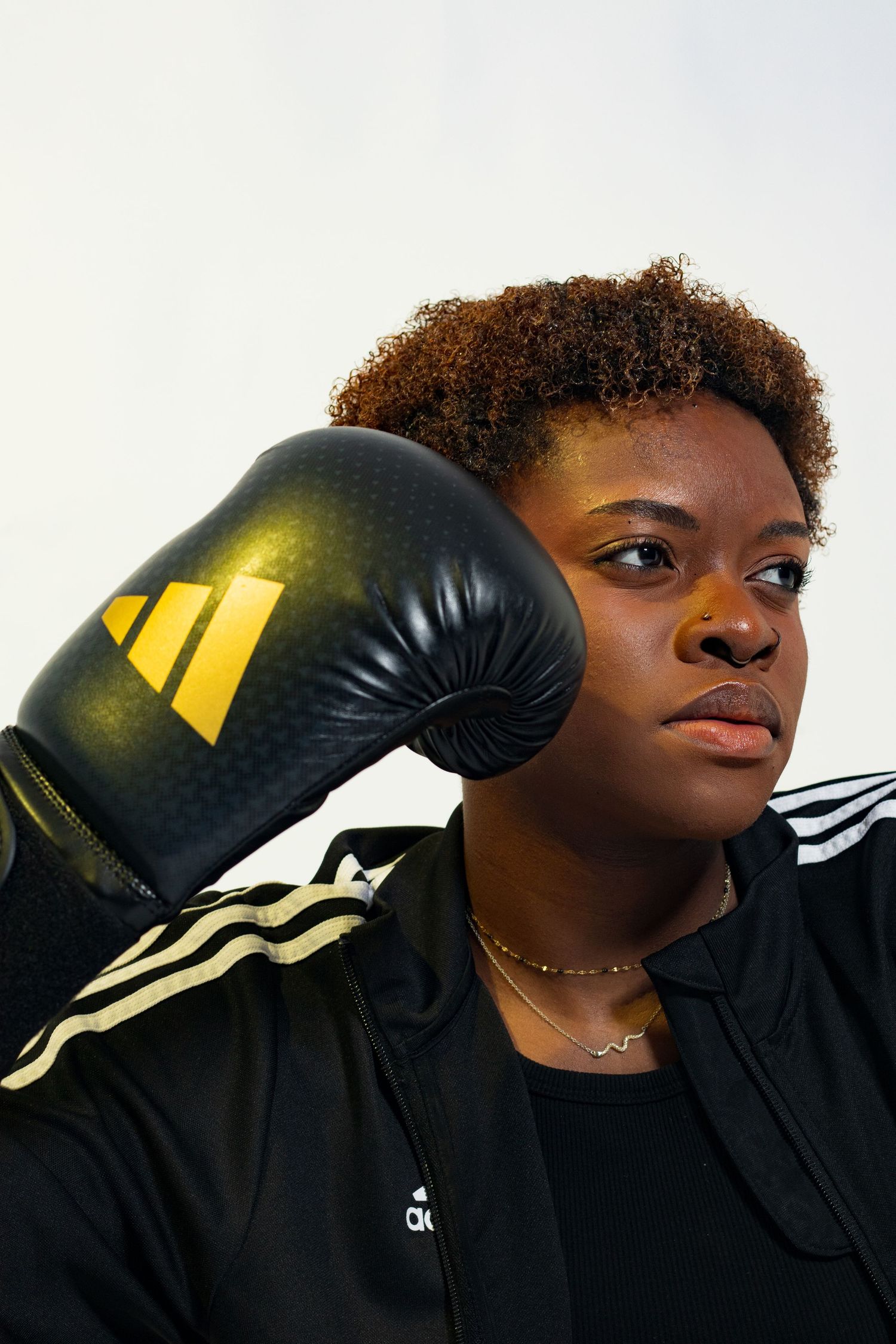 Young Black female boxer in Adidas jacket holding a black and gold boxing glove beside her head.