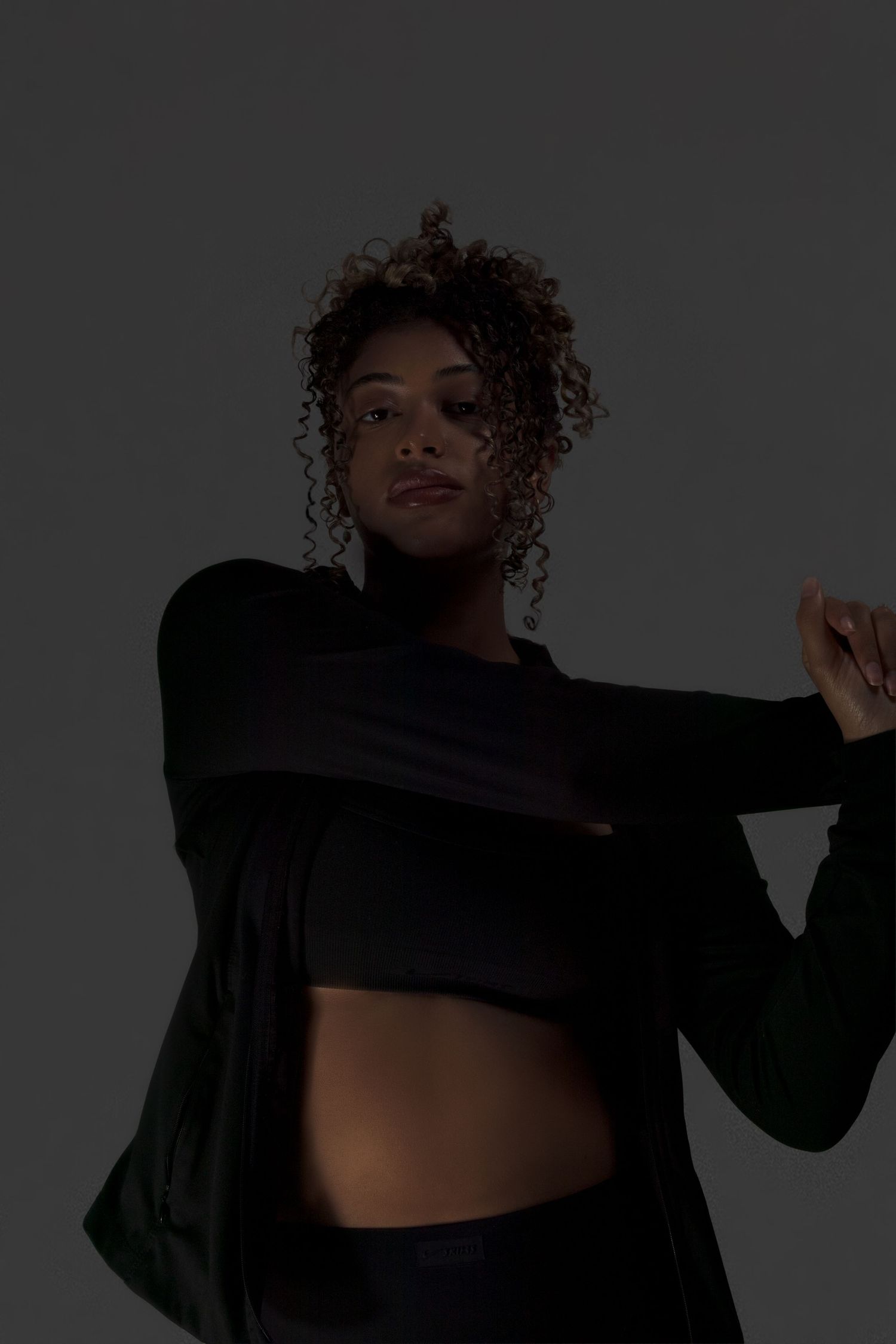 A woman in a black crop top and jacket poses dynamically against a dark gray studio background.