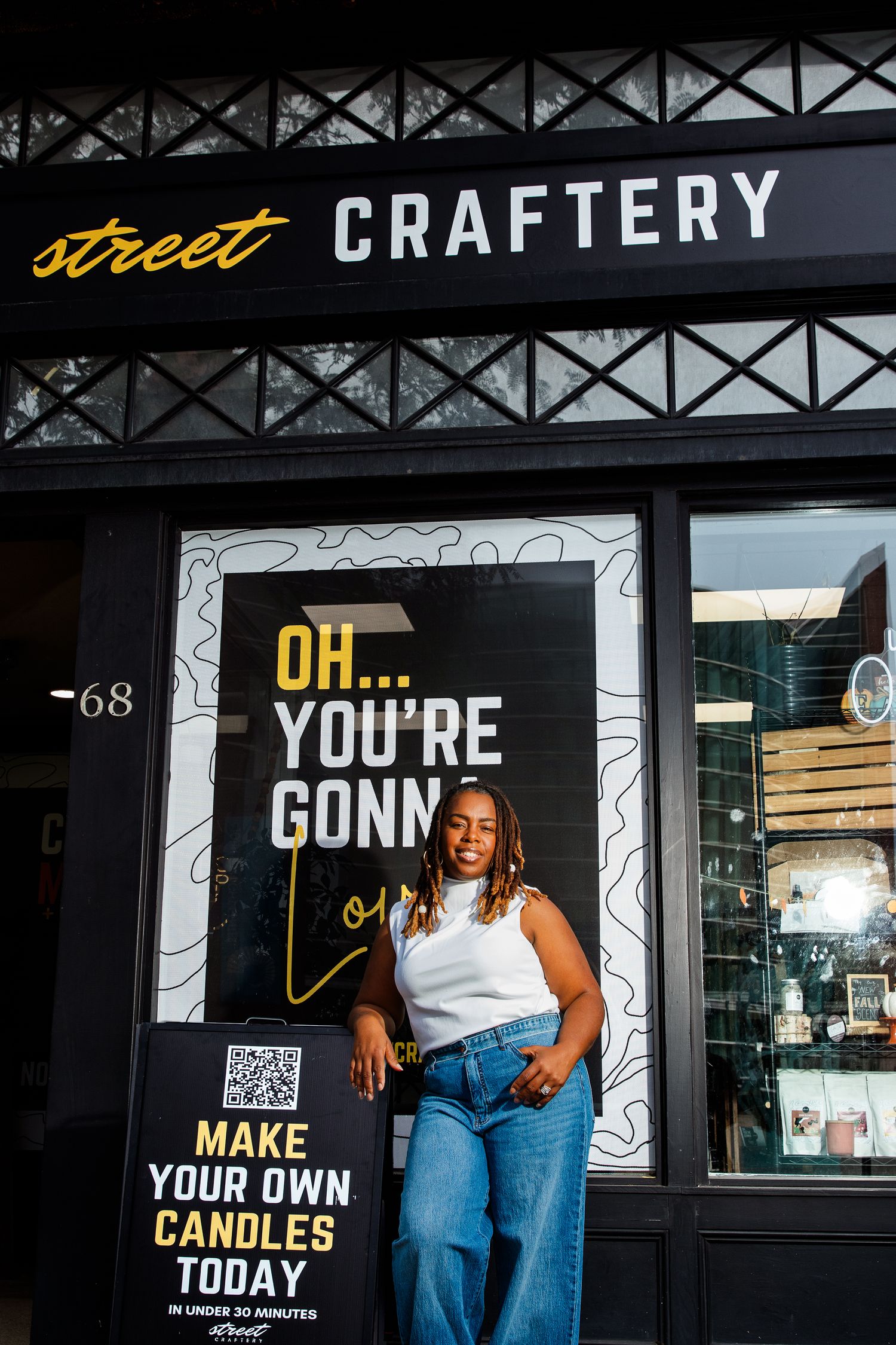Woman in white top and jeans leaning against Street Craftery storefront with make your own candles sign.