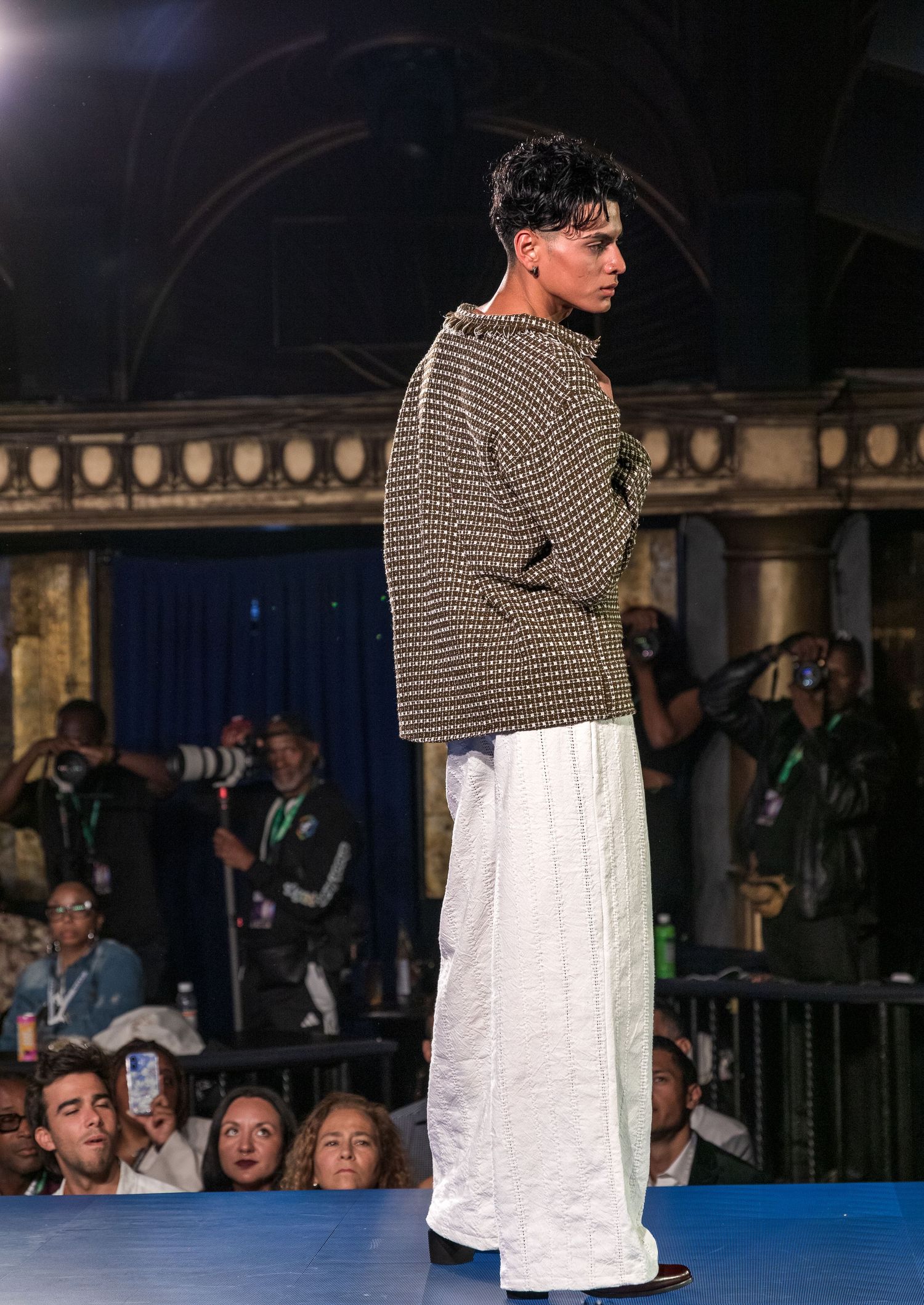 Male model on runway wearing textured knit sweater and wide-leg white trousers at evening fashion show.