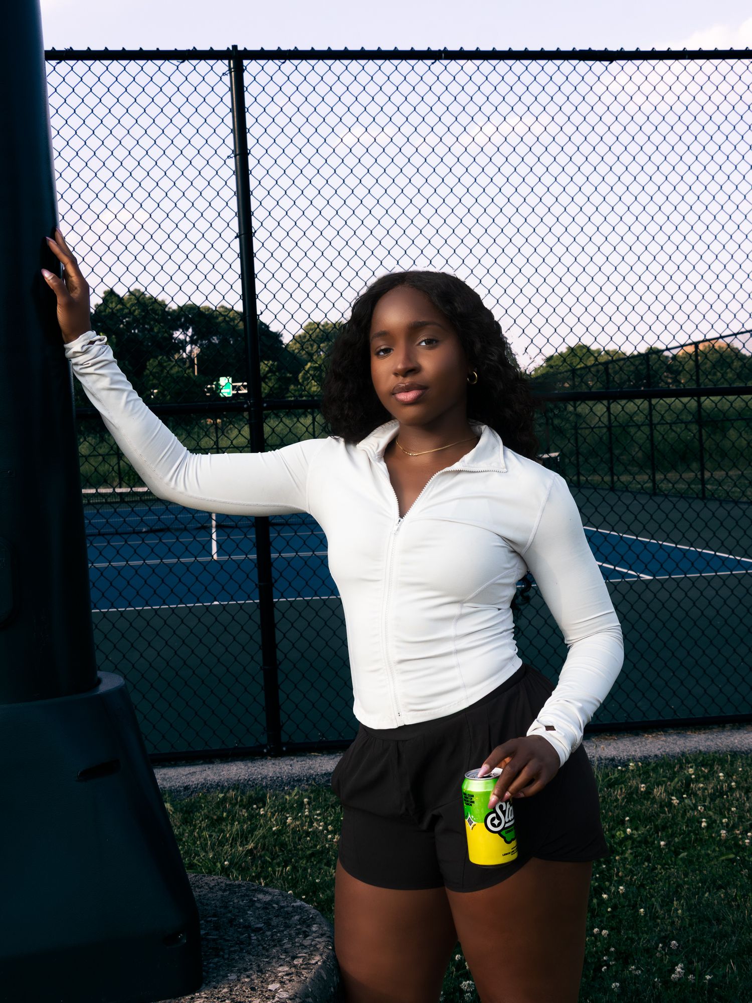 Young woman leaning against a fence at a tennis court holding a small green soda can at dusk.