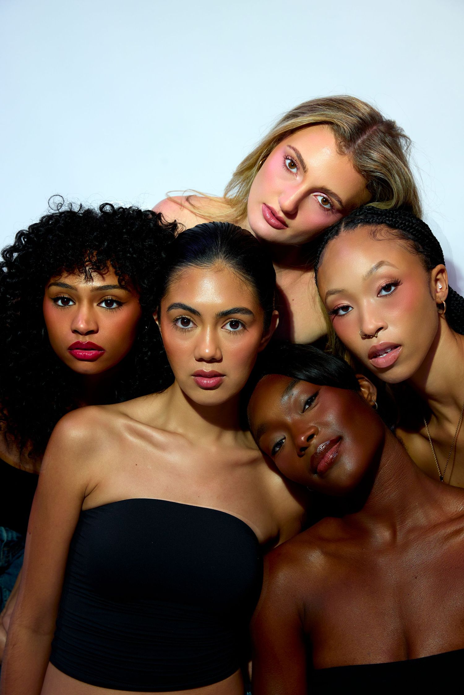 Five diverse women with glowing makeup pose together against a white background in a beauty editorial shot.