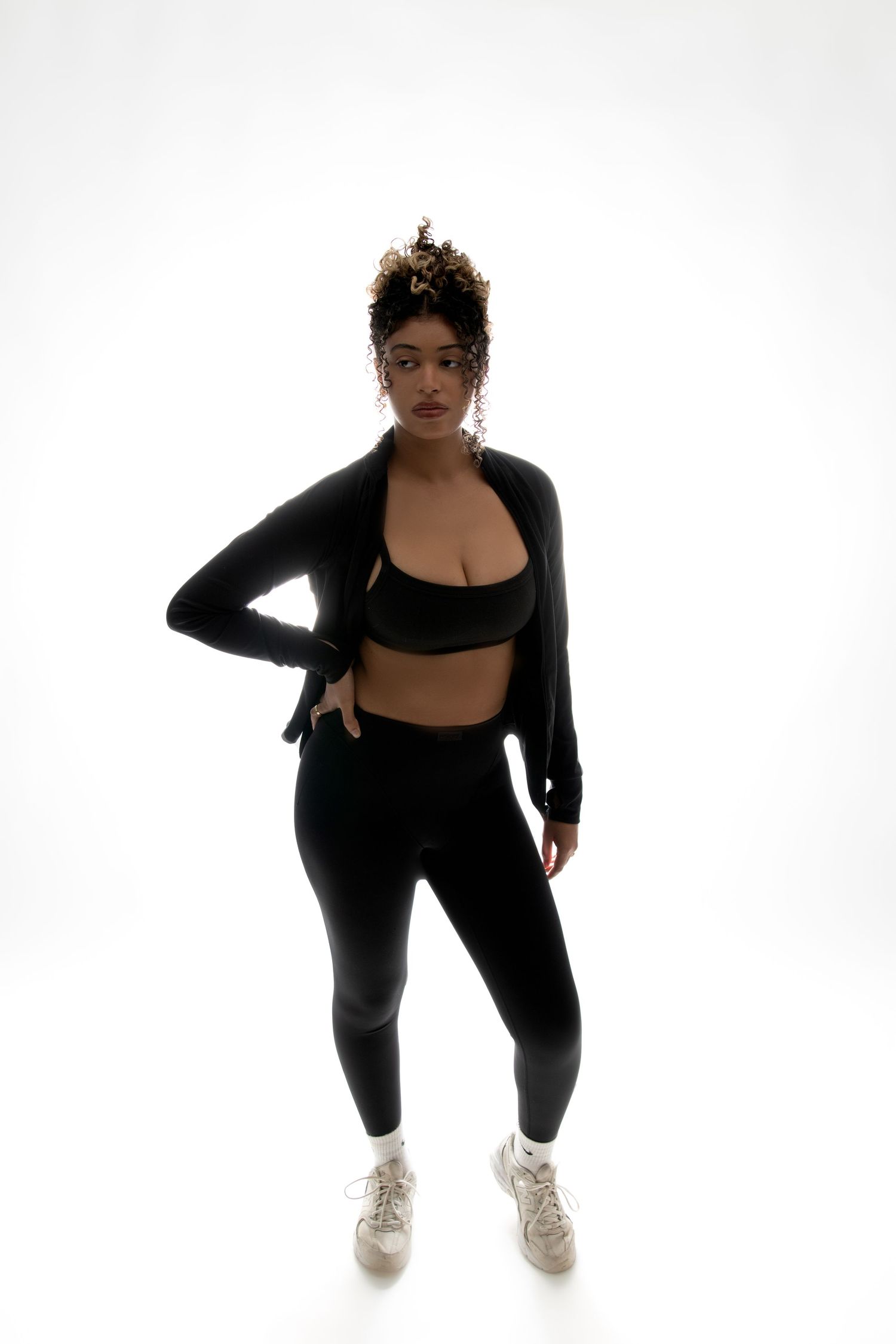 A woman in a black sports bra, jacket, and leggings poses confidently against a white background.