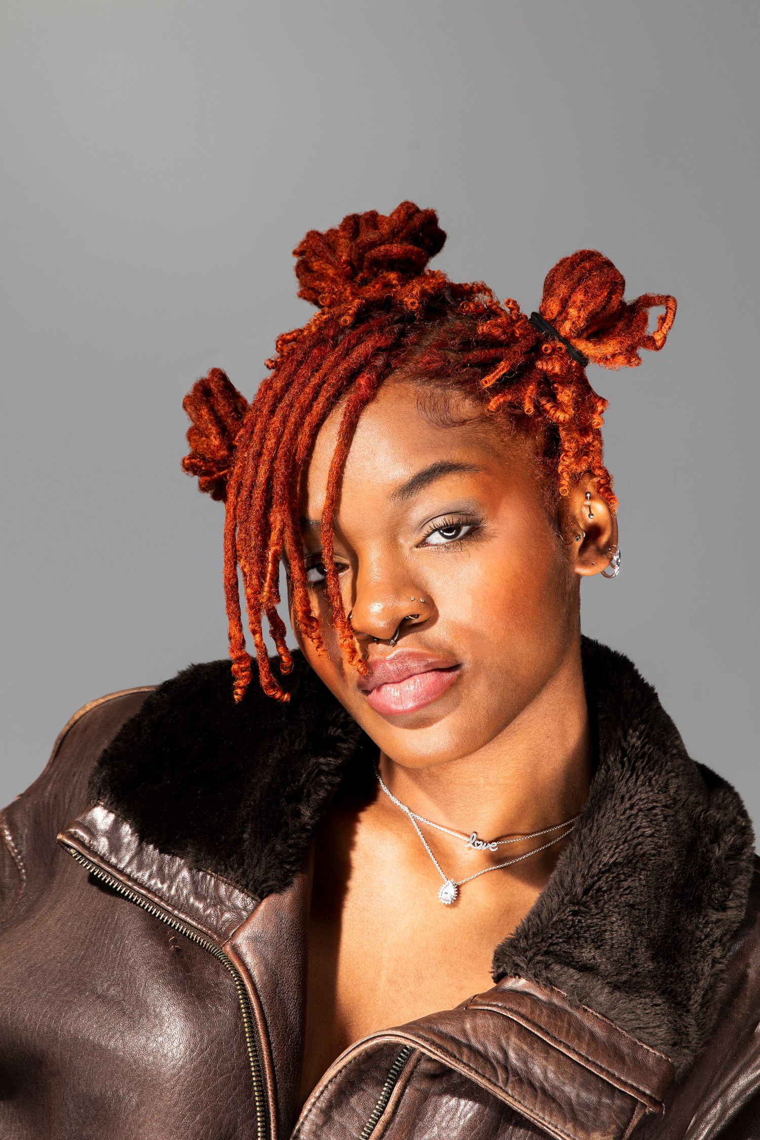 Close-up portrait of woman with vibrant orange dreadlocks wearing brown leather jacket with fur collar.