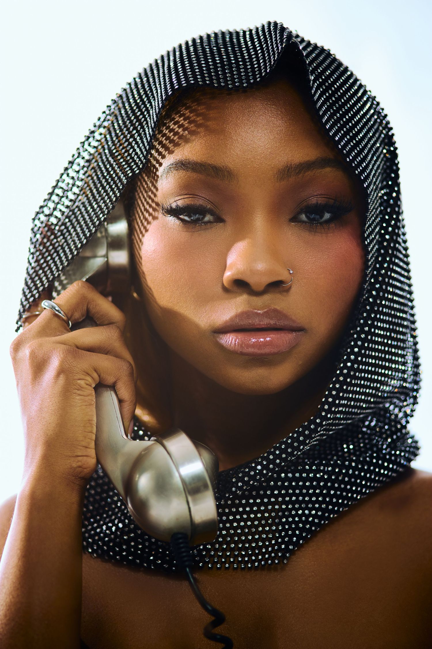 Young Black woman in a metallic dotted hood holding a retro silver telephone handset near her face.