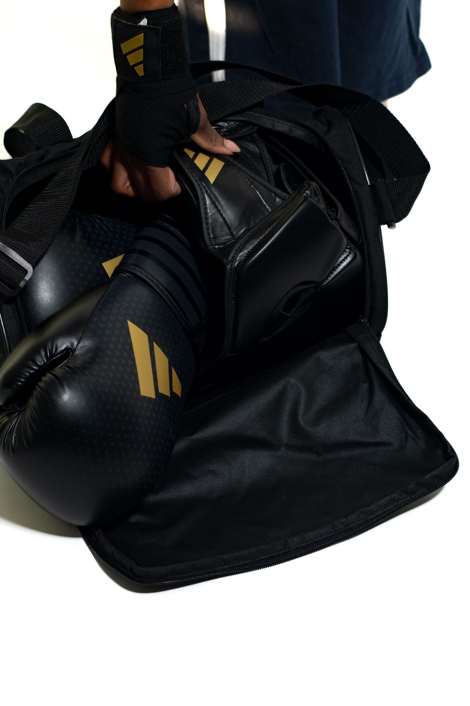 Black and gold Adidas boxing gloves and gear packed in open sports duffel bag on white background.