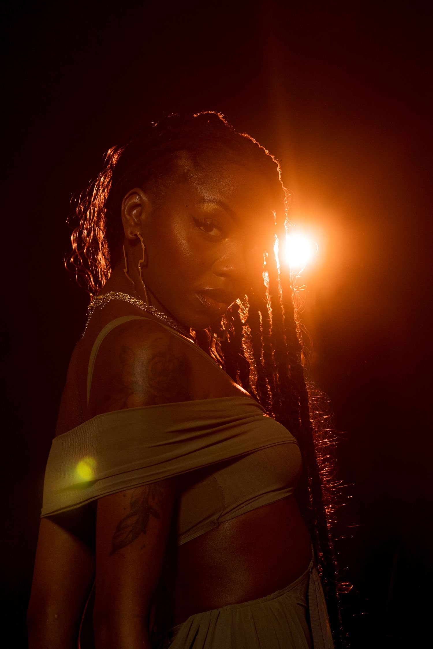 A Black woman with long braids poses in a golden olive dress under dramatic orange backlight in a dark studio.