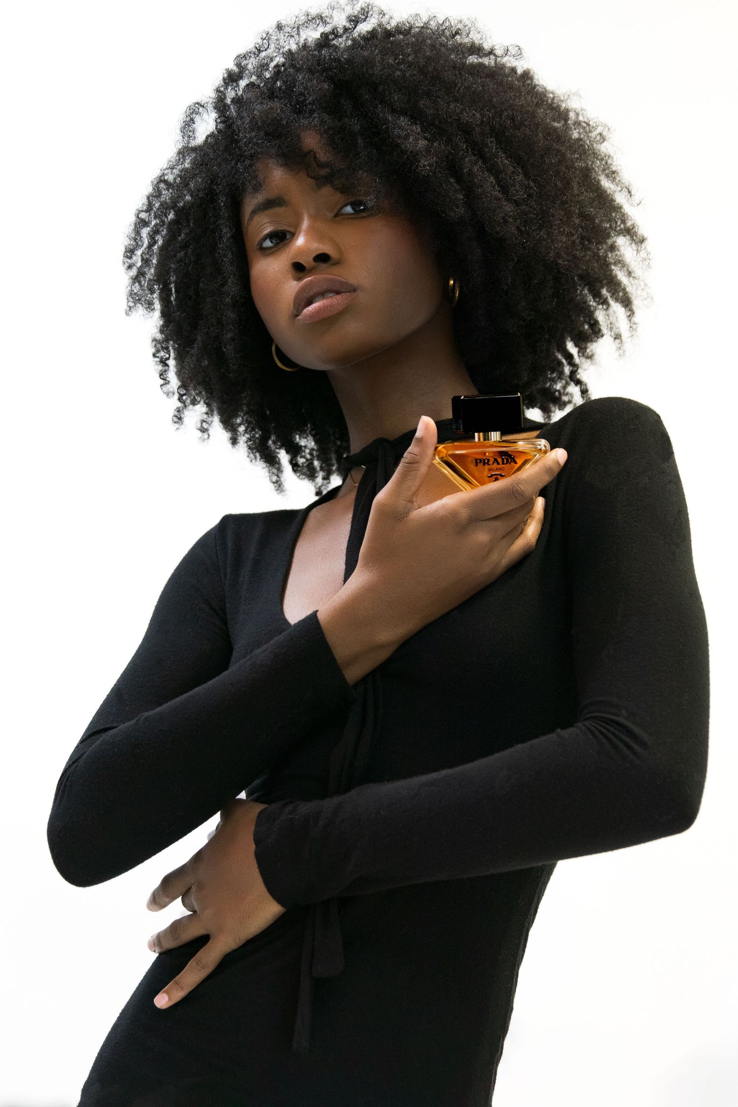 Woman with natural afro hair holding a Prada perfume bottle against her chest in a black dress.