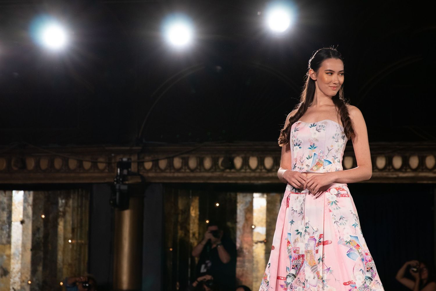 Model wears pink floral strapless ball gown on elegantly lit fashion runway.