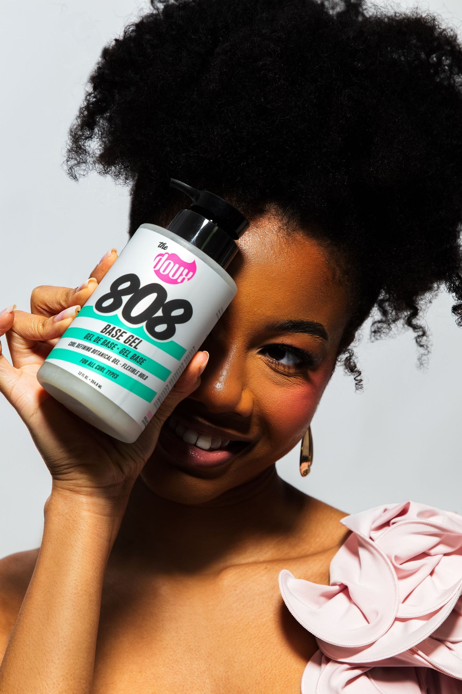 Smiling woman holding The Doux 808 Base Gel jar beside her face with natural curly hair.