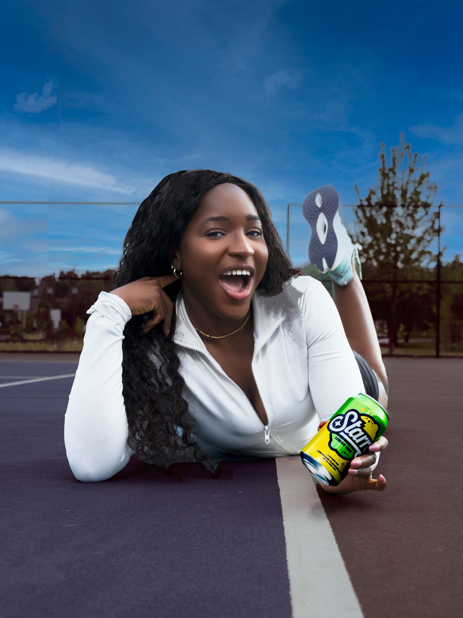 Cheerful young woman lying on a tennis court holding a Starry soda can with a blue sky background.