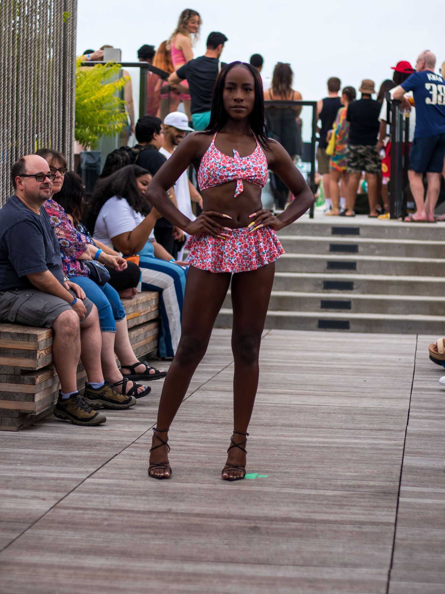 Tall slender model in floral bikini top and mini skirt walks outdoor boardwalk fashion show runway.