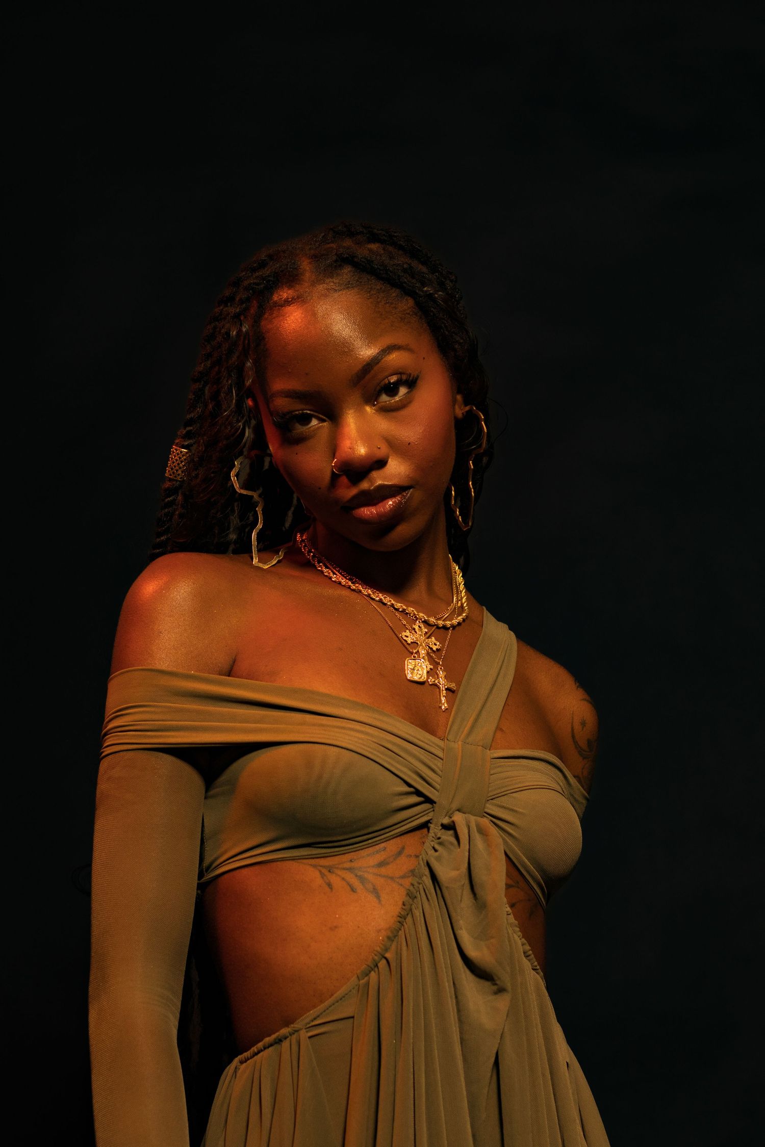 Black woman in olive off-shoulder crop top and skirt with gold cross necklace posing against black background.