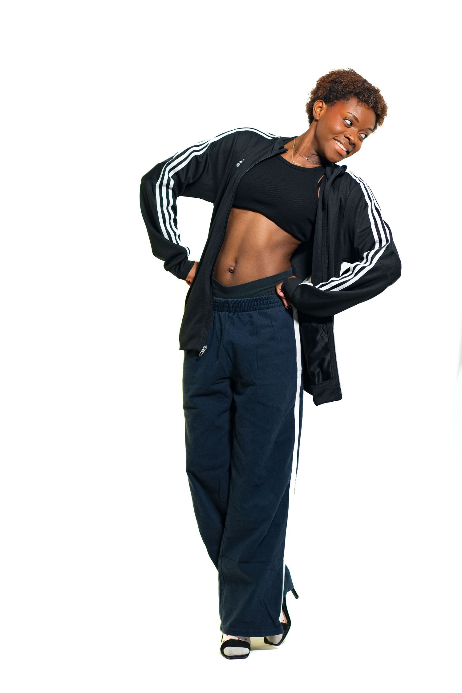 Athletic Black woman posing in Adidas tracksuit with crop top showing toned abs on white background.