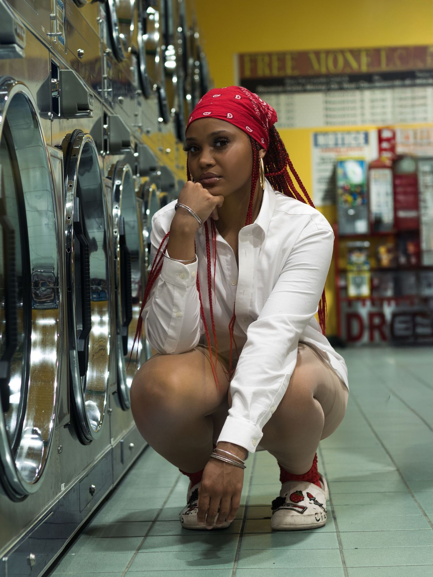 Woman with red braids and red bandana crouches by washing machines in laundromat.