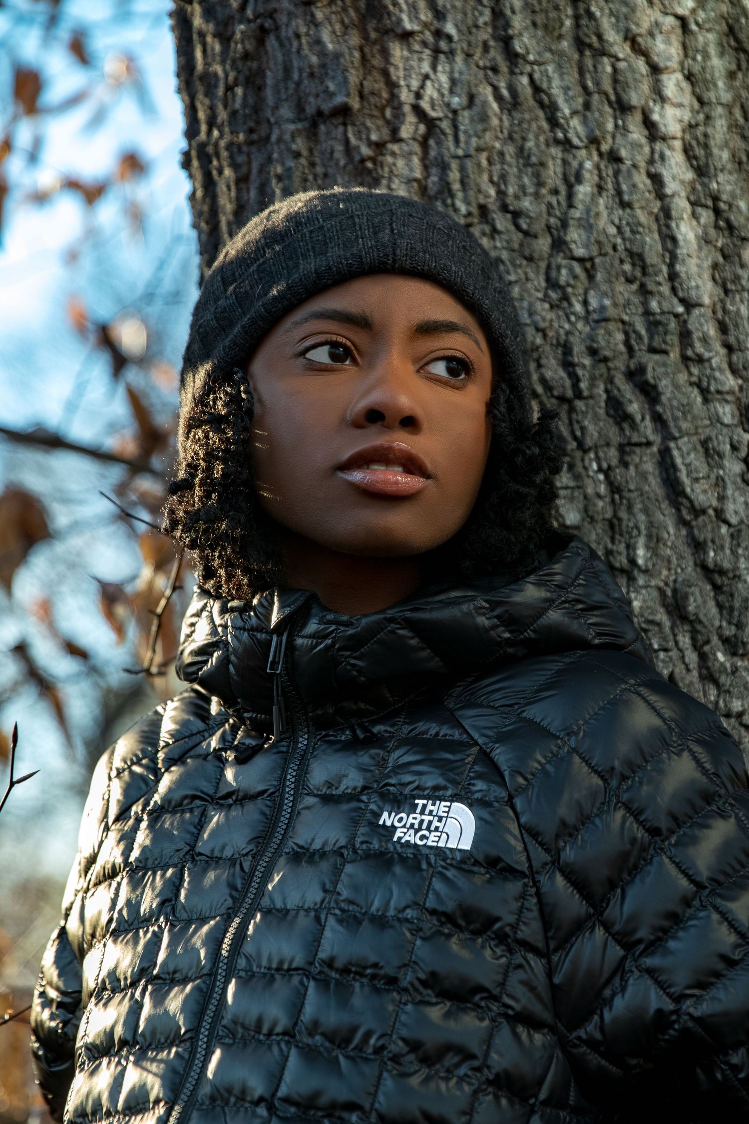 Young woman in North Face black puffer jacket and grey beanie leaning against tree outdoors.