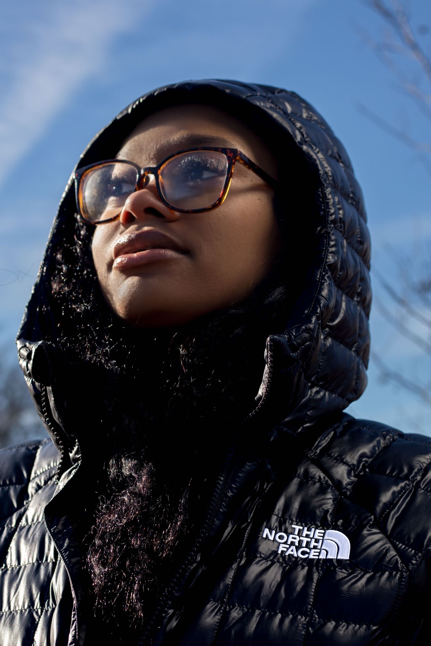 Woman wearing glasses and black North Face hooded puffer jacket looking up at winter sky.