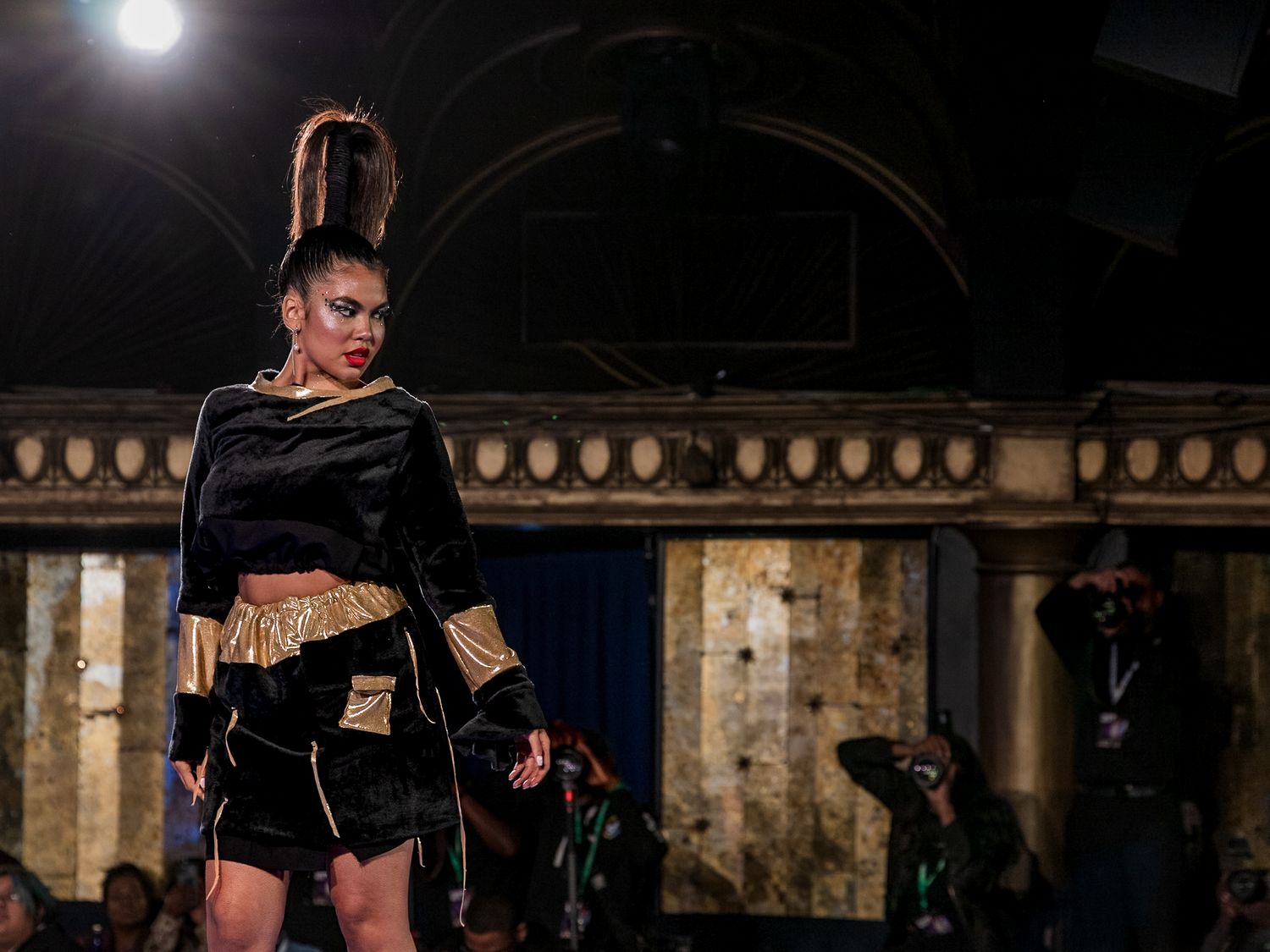 Model walks runway in black and gold crop top and mini skirt ensemble at fashion show.