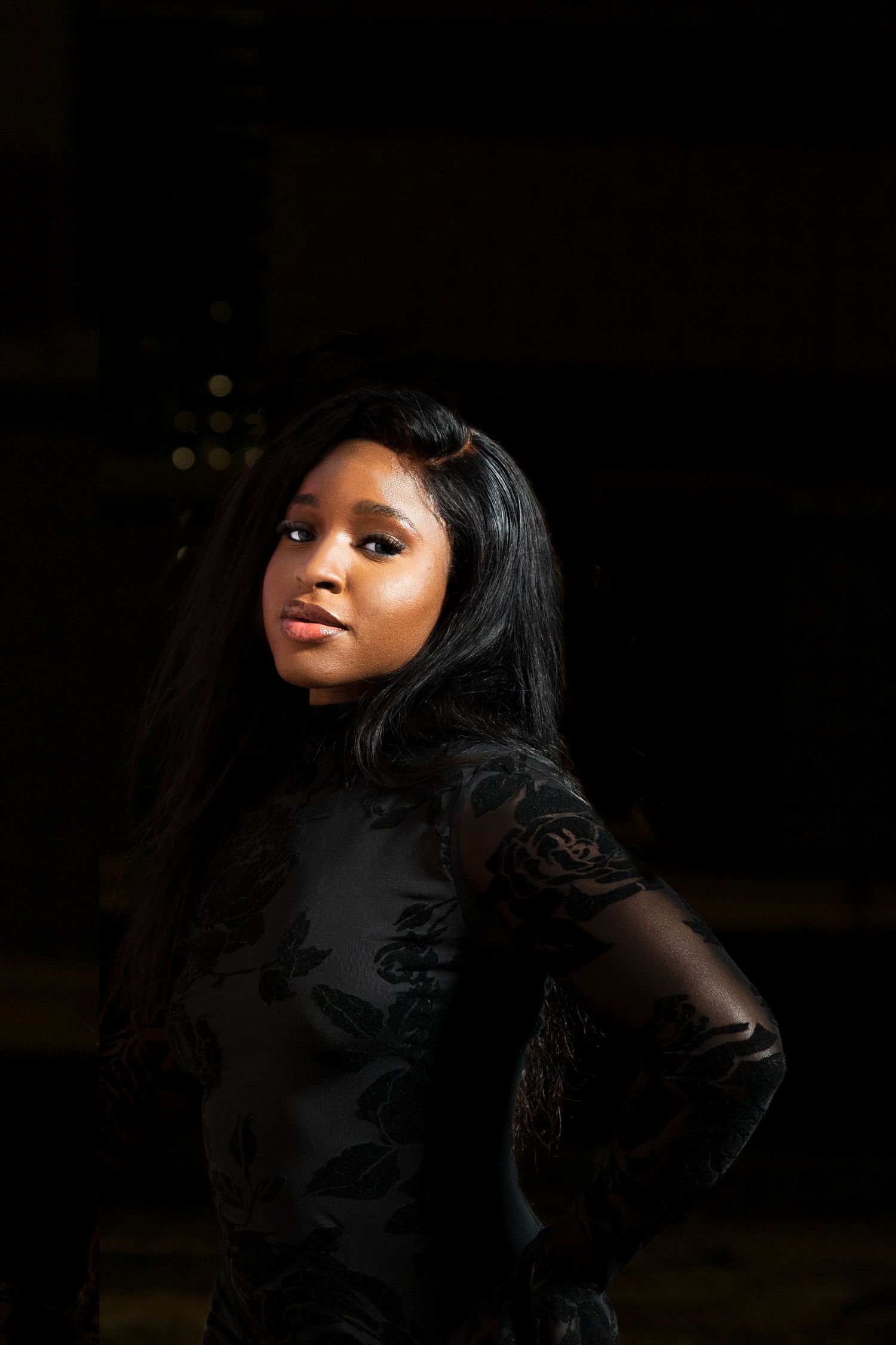 Elegant Black woman in a black floral mesh dress posing with dramatic shadow lighting.