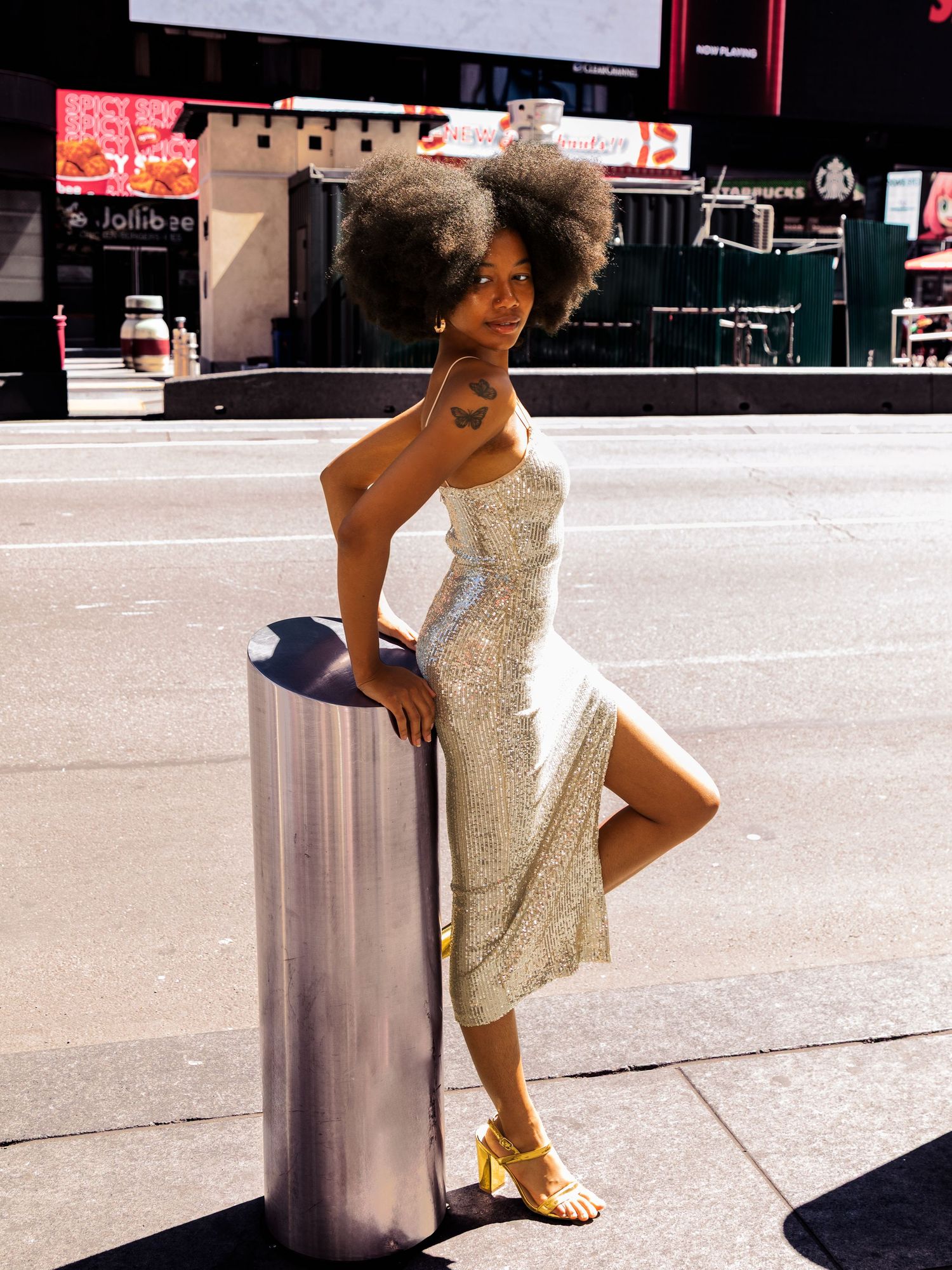 Stylish woman in sequin midi dress with afro hair leaning on silver bollard in Times Square NYC.