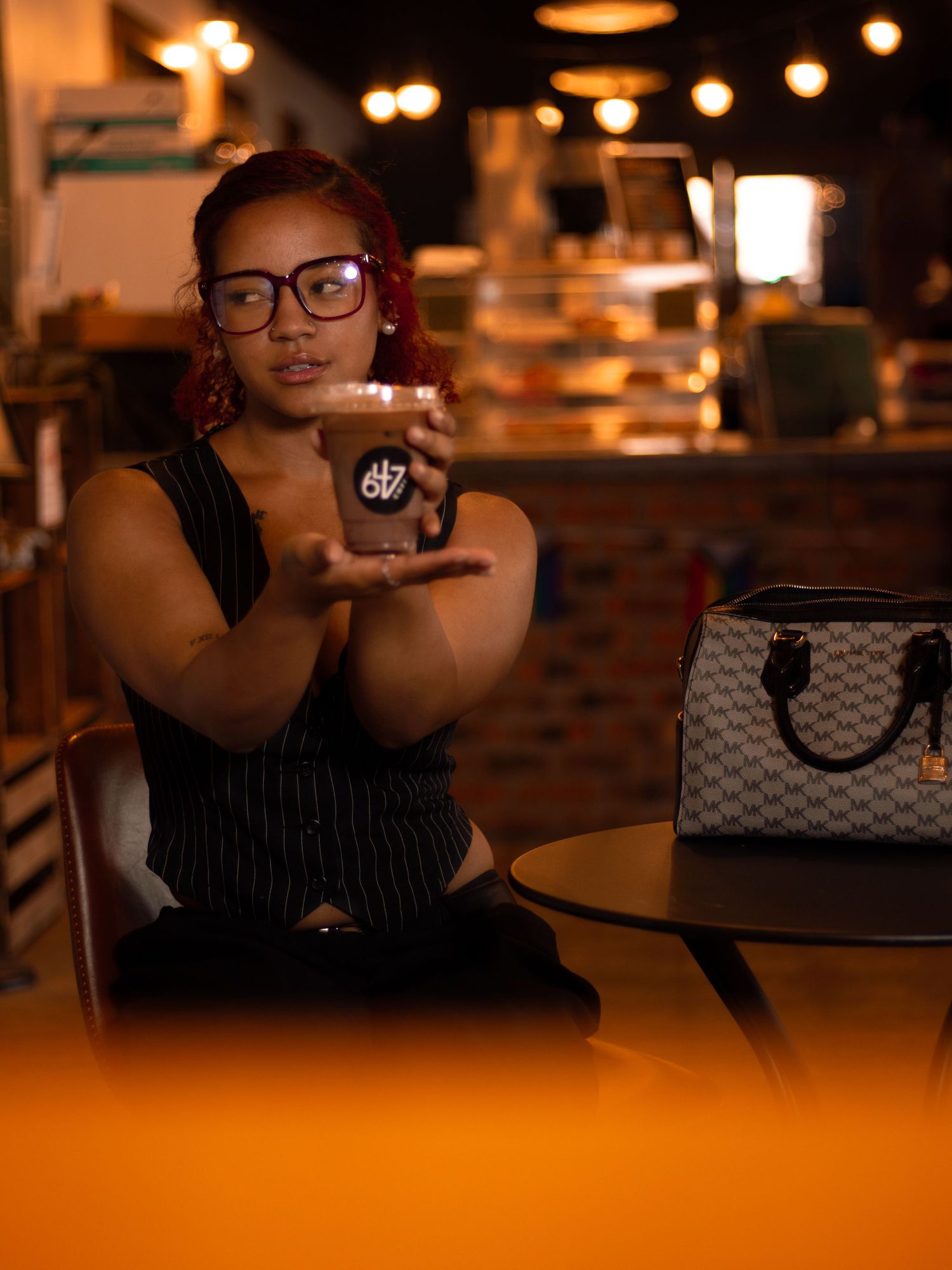 Woman with red hair and glasses holding iced coffee cup beside designer bag in cafe.
