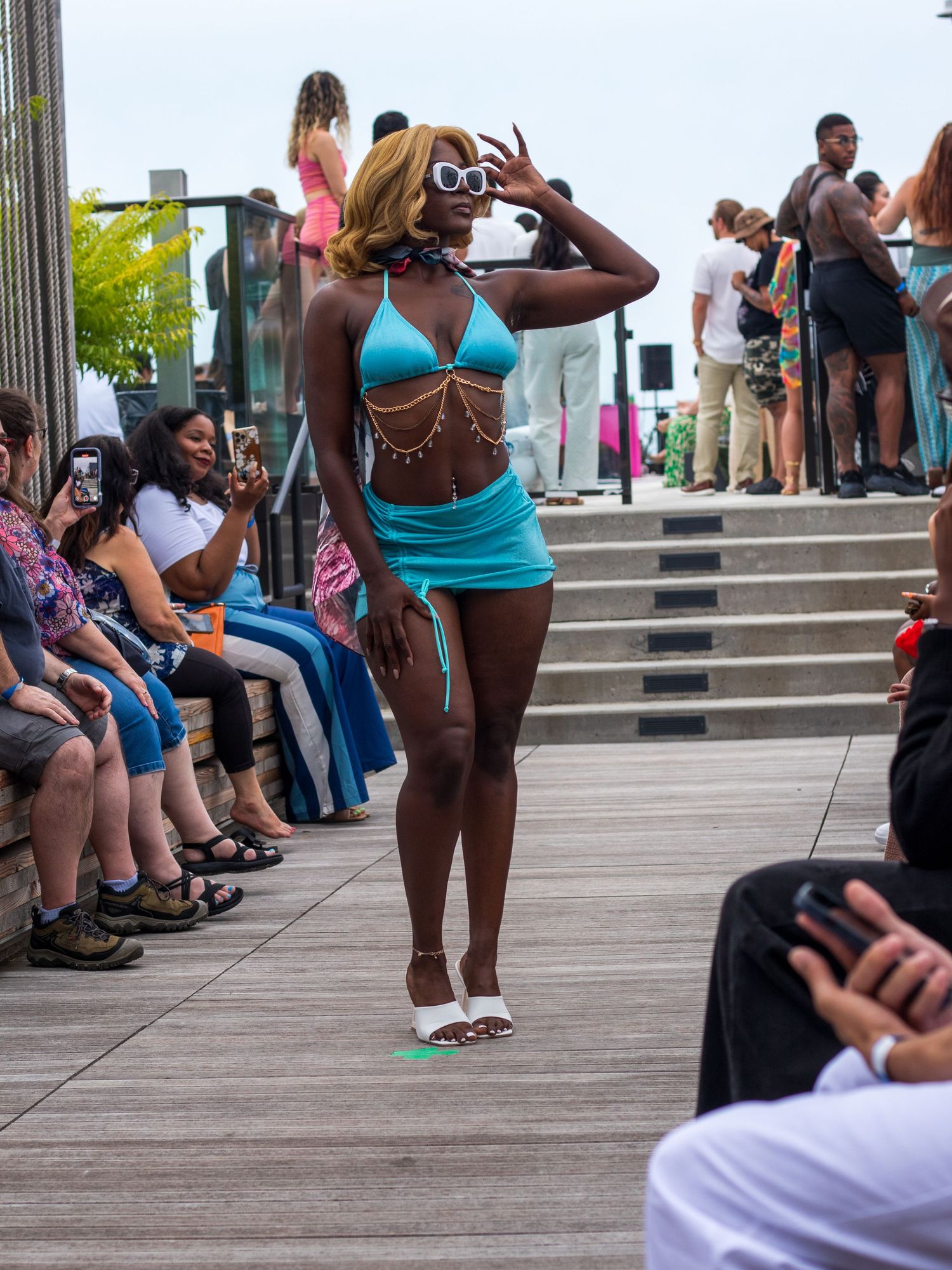 Model in turquoise bikini set with gold chains and white sunglasses on outdoor runway.
