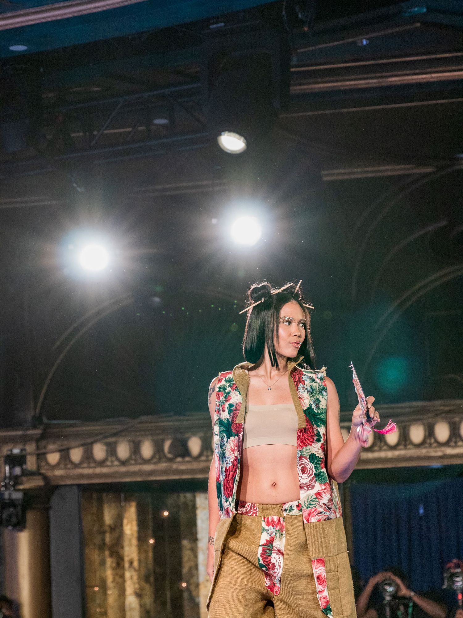 Model struts runway in floral vest with crop top and tan pants at indoor fashion show.