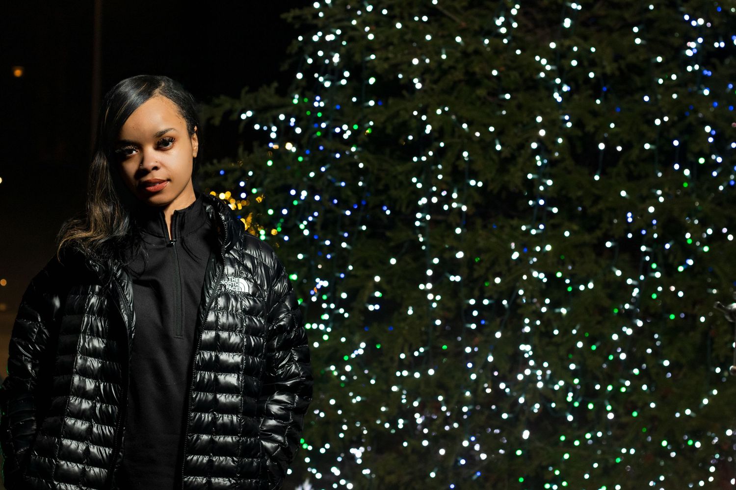 Woman in black puffer jacket standing before illuminated Christmas tree at night.