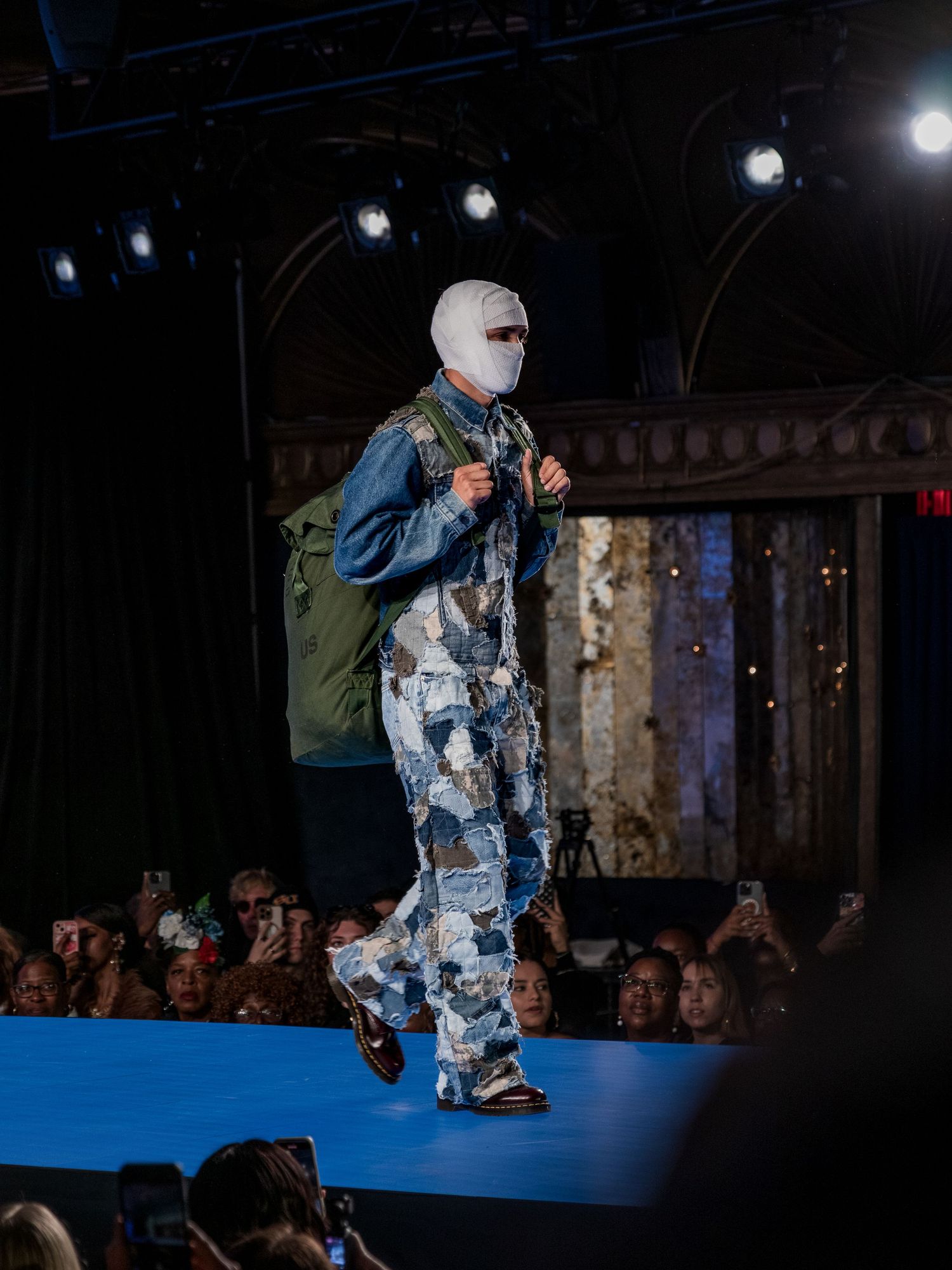 Male model walks runway in bandage-wrapped face, patchwork denim outfit and green backpack.