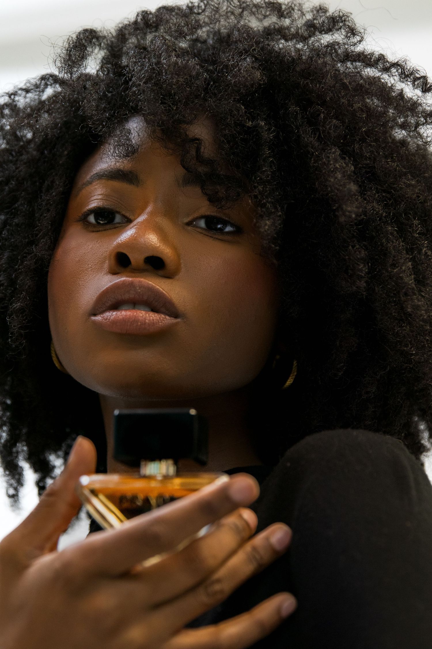 Close-up portrait of a woman with afro hair holding a Prada perfume bottle near her face.