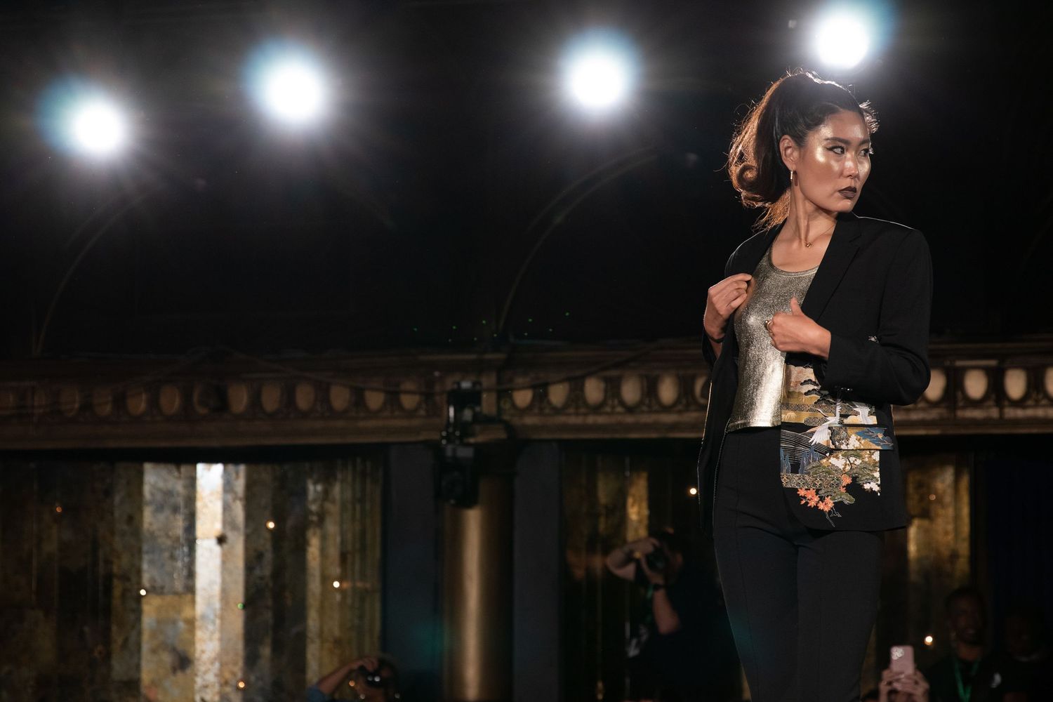 Model walks runway in all-black ensemble carrying metallic clutch at fashion show.