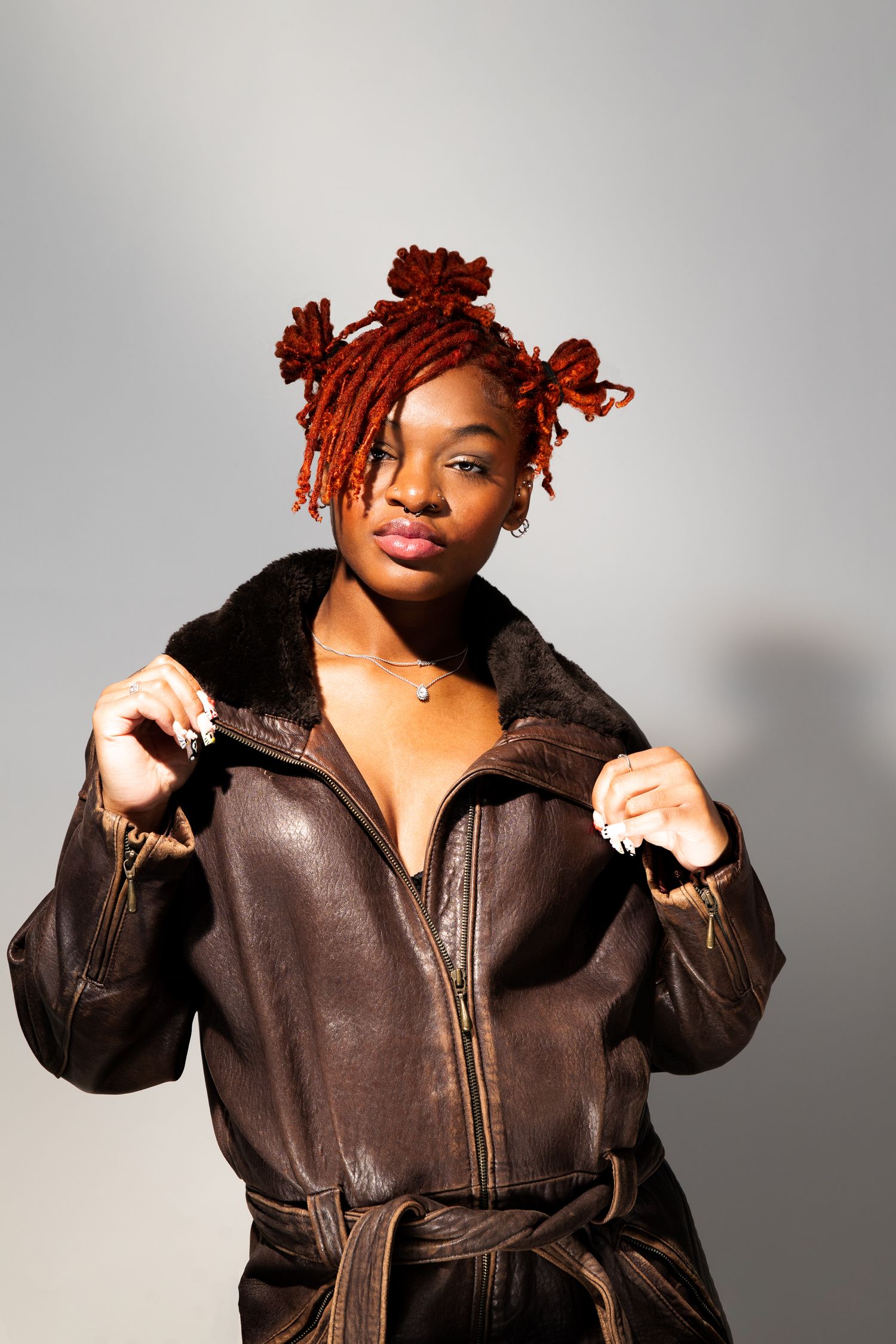 Fashion model with orange locs adjusts brown vintage leather belted jacket in studio portrait.