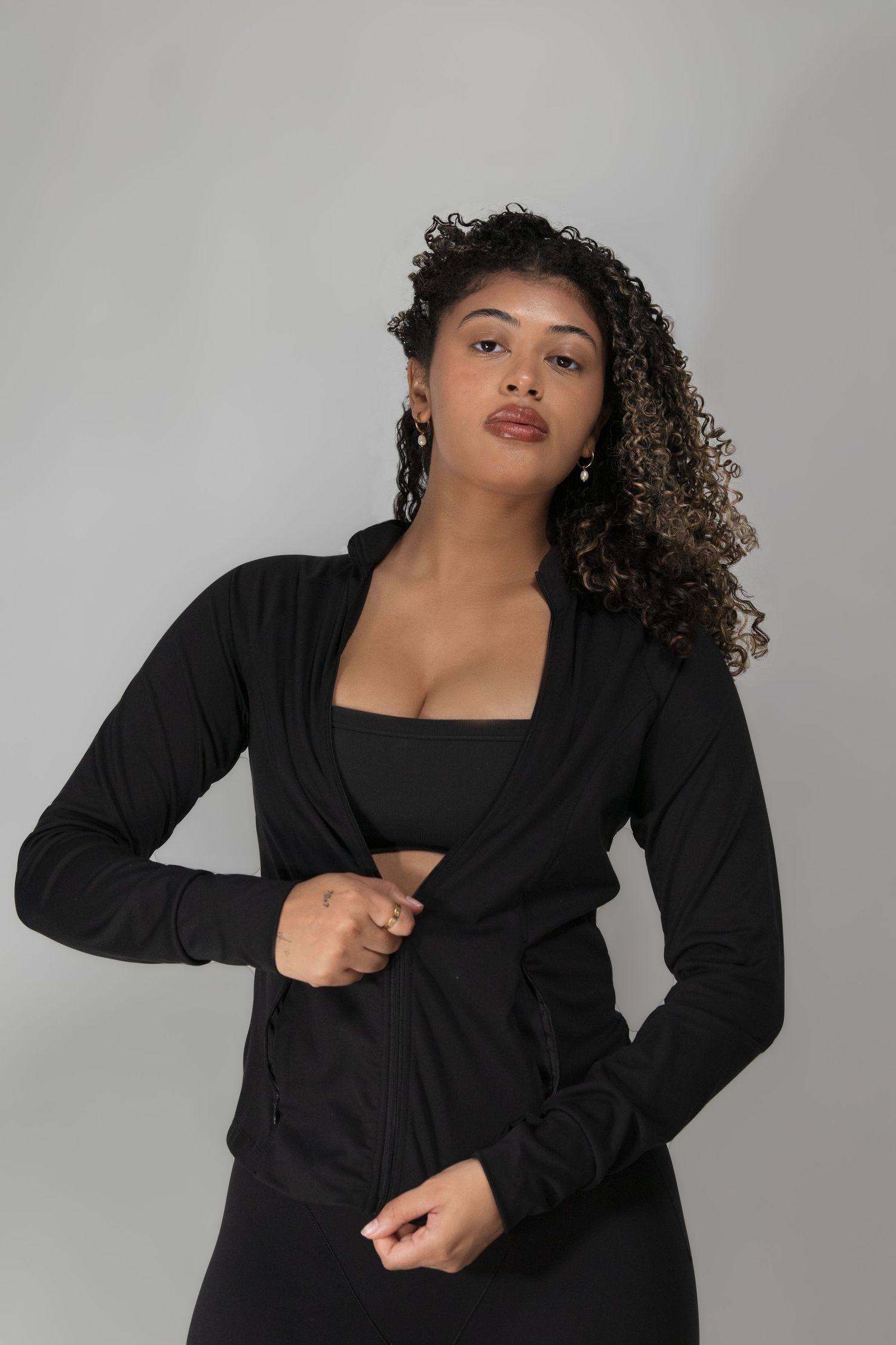 A woman with curly highlighted hair adjusts her black zip-up jacket in a studio portrait.
