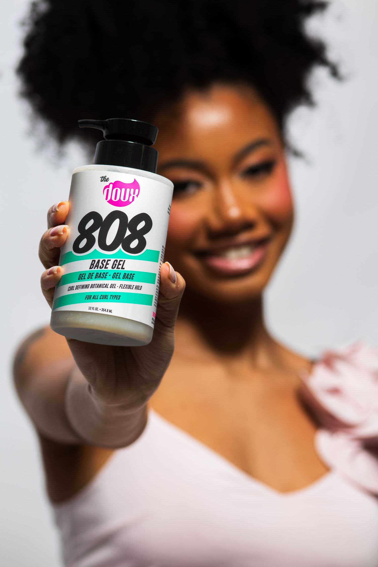 Smiling woman with afro hair holding The Doux 808 Base Gel jar extended toward camera.