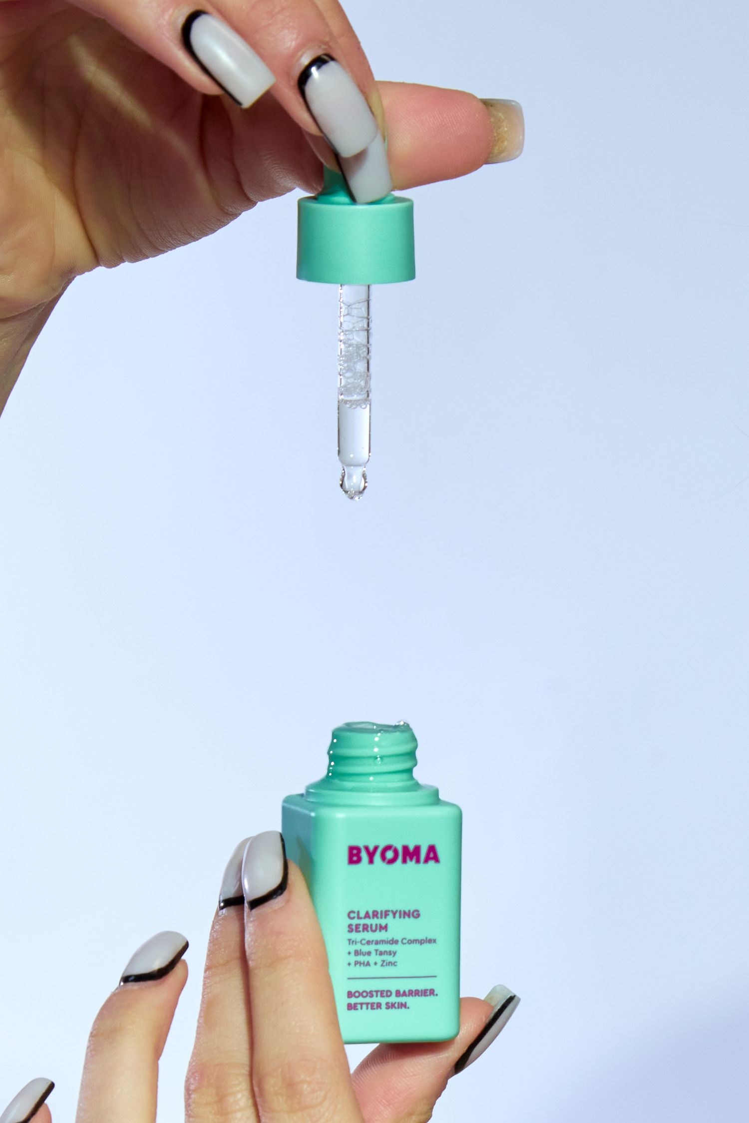 Hand holding Byoma Clarifying Serum dropper bottle with teal cap, dispensing a drop of serum on white background.
