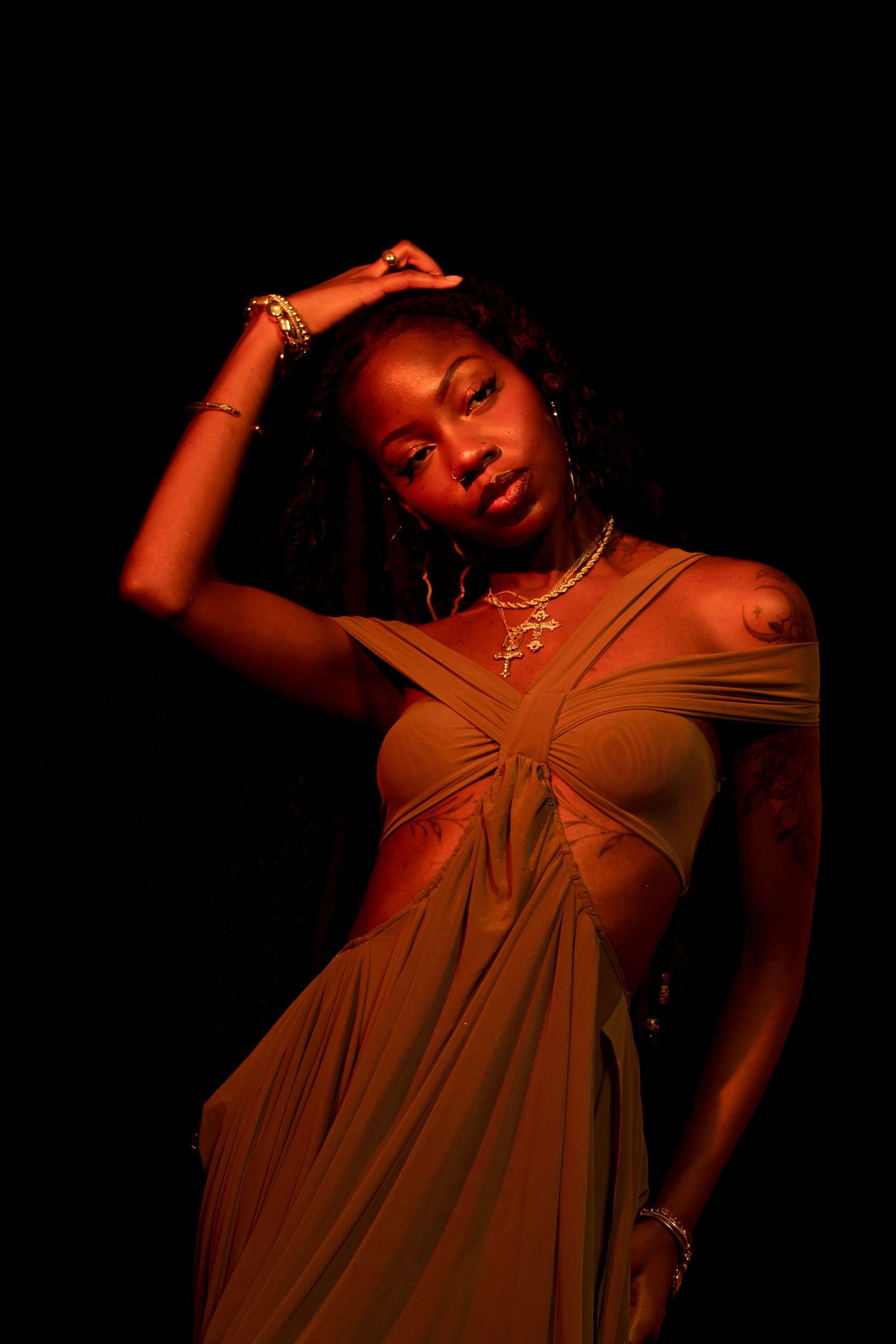 A Black woman in an olive wrap dress poses with hand on head, adorned with gold jewelry in warm orange studio light.