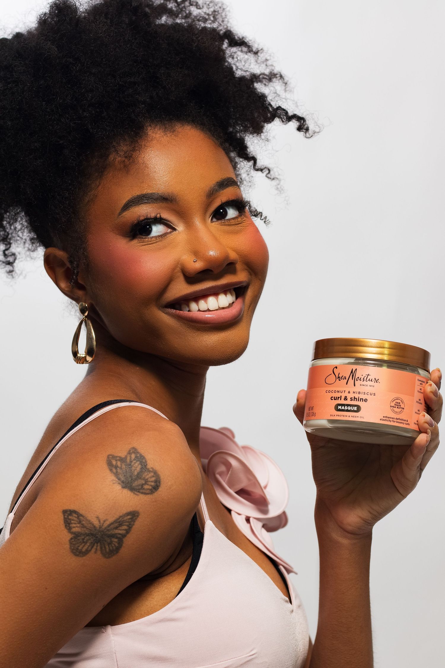 Smiling woman with afro hair holding SheaMoisture Curl & Shine mousse jar, butterfly tattoo visible.