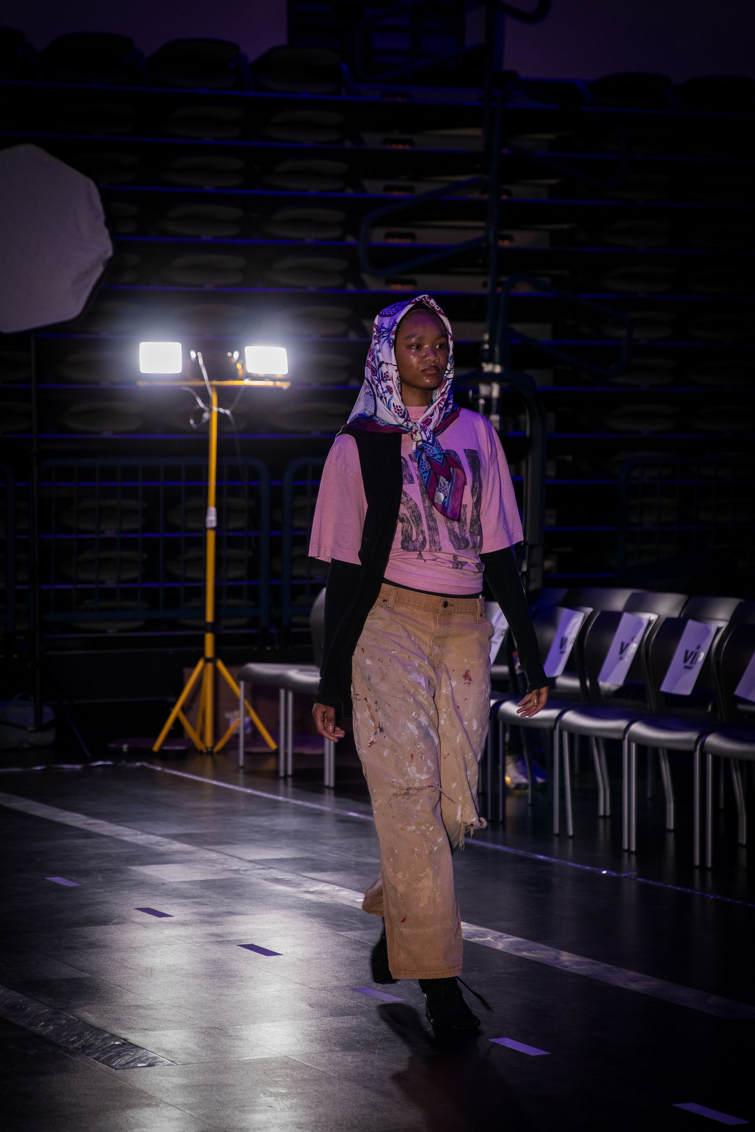 A model walks the runway in a purple graphic hoodie and tan cargo pants at an indoor fashion show.