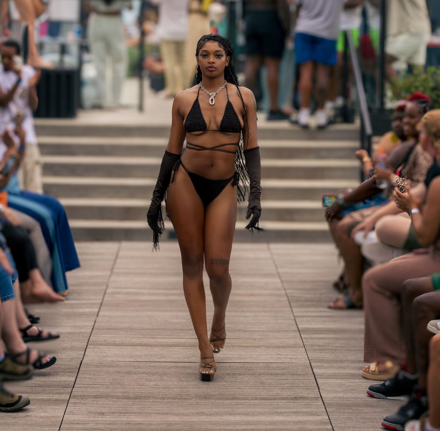 Model in black string bikini with gloves and necklace walks outdoor fashion runway.