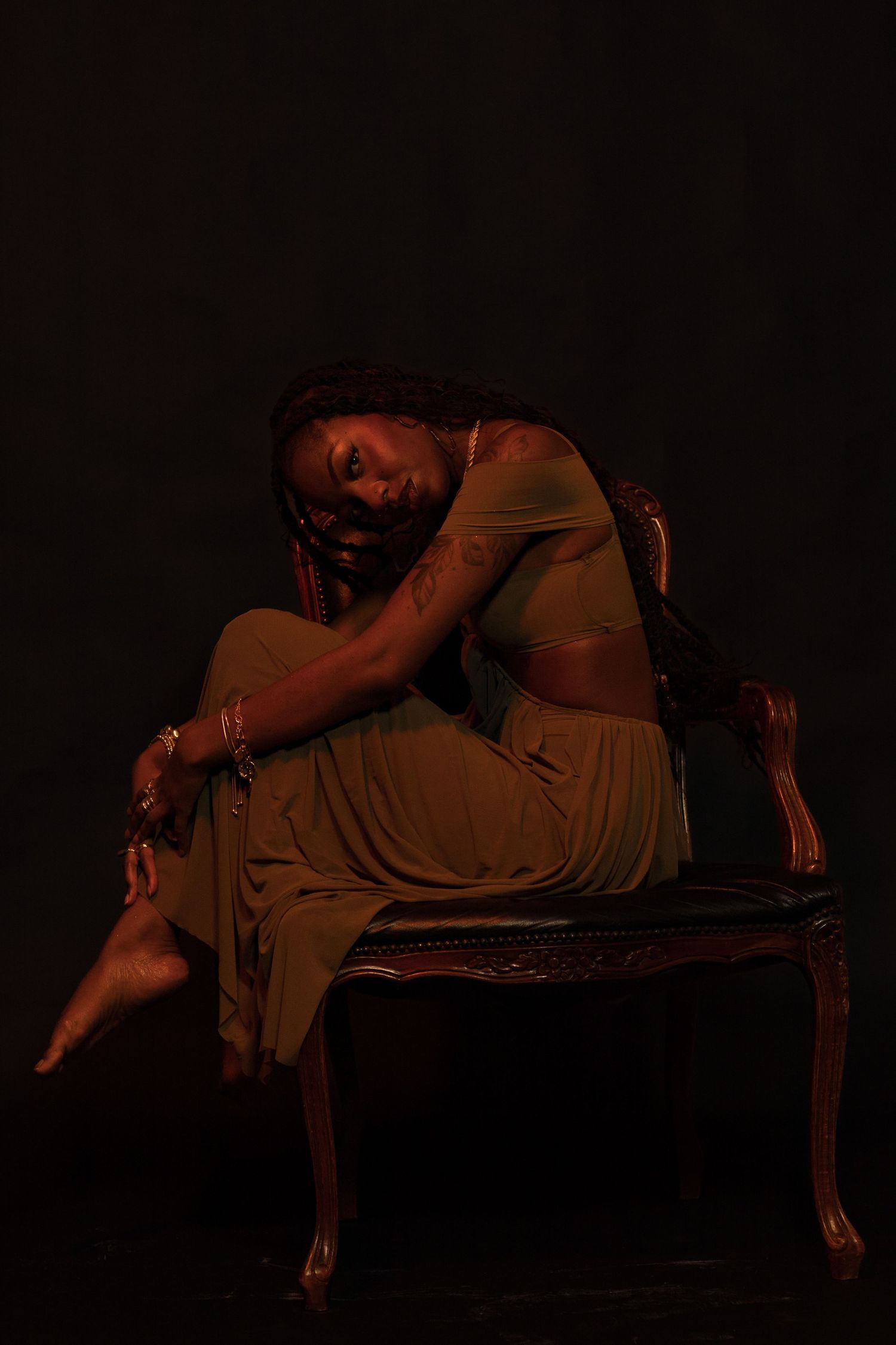 A Black woman in an olive dress sits curled on an antique chair in a moody dark studio with warm orange lighting.