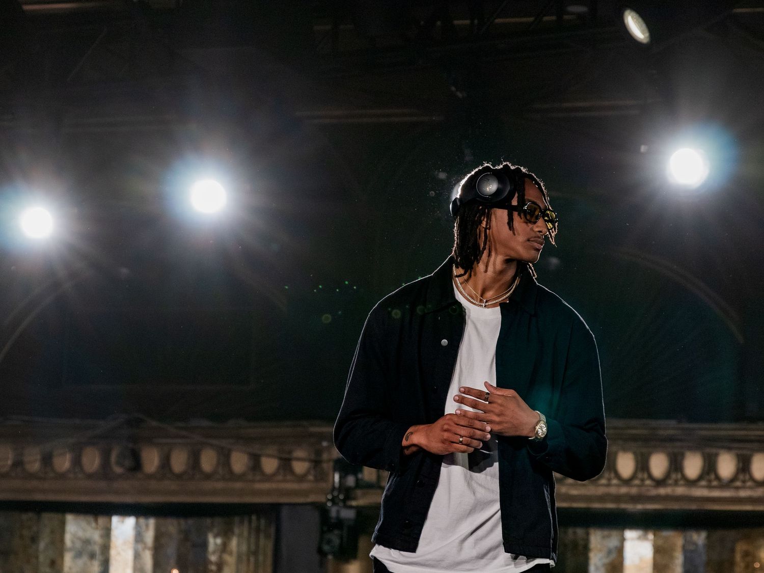Male performer in dark jacket and headphones stands under stage lights at event.