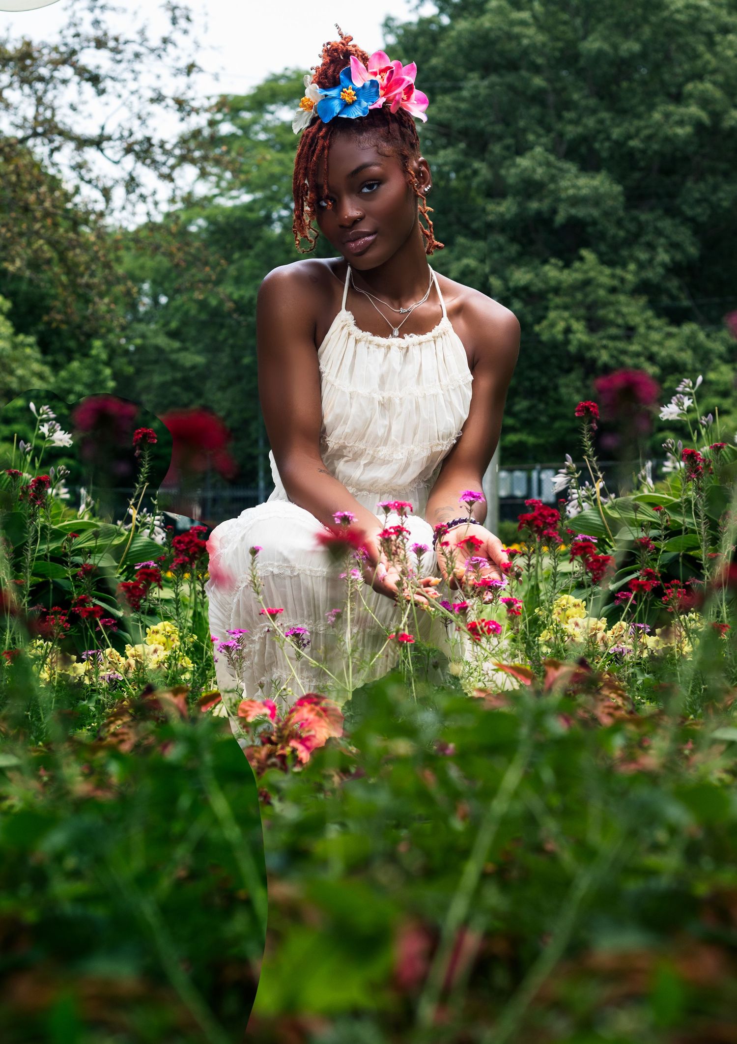 Woman in white dress with colorful floral headpiece kneels among vibrant garden flowers outdoors.