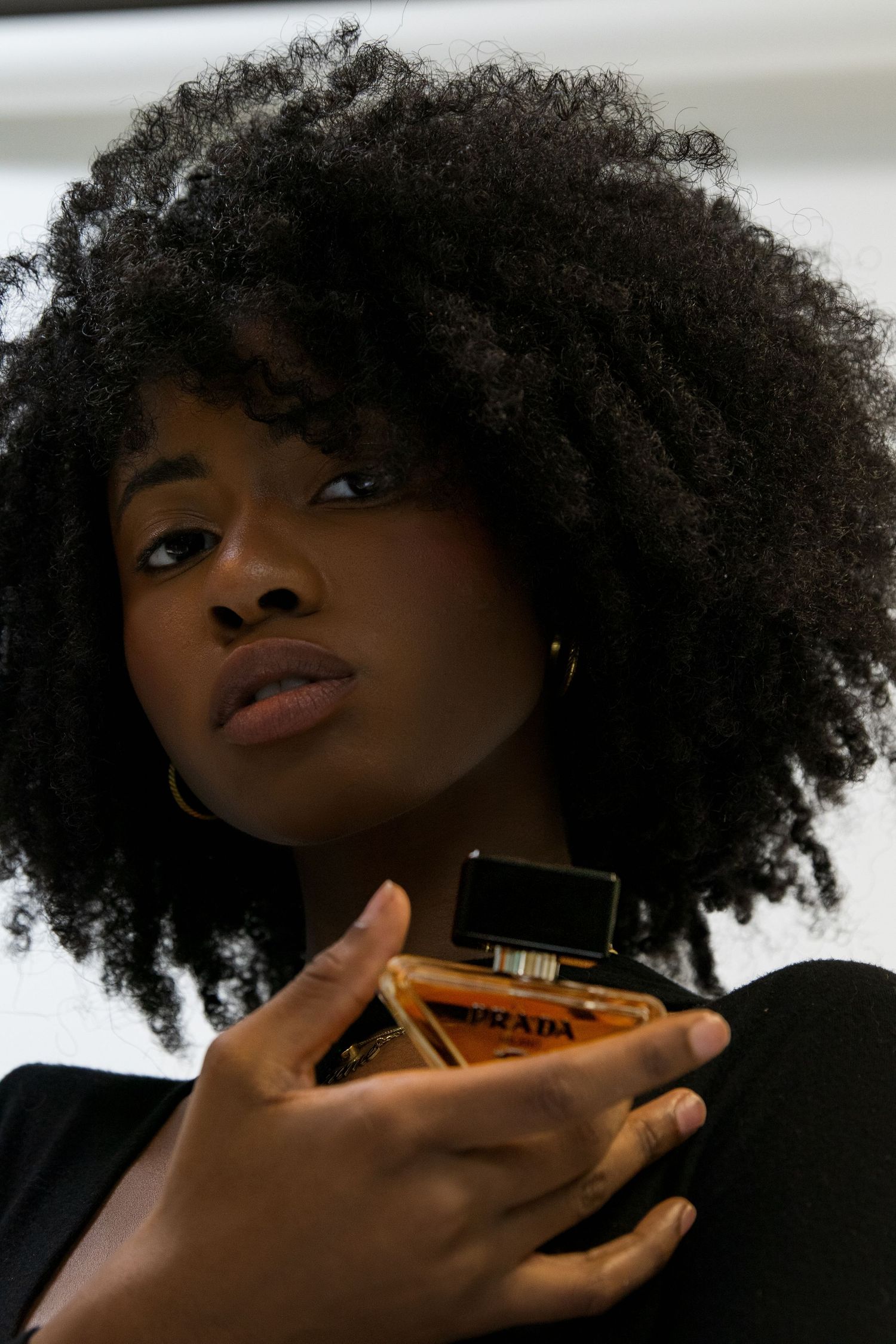Close-up of a woman with voluminous afro hair holding a Prada fragrance bottle confidently.
