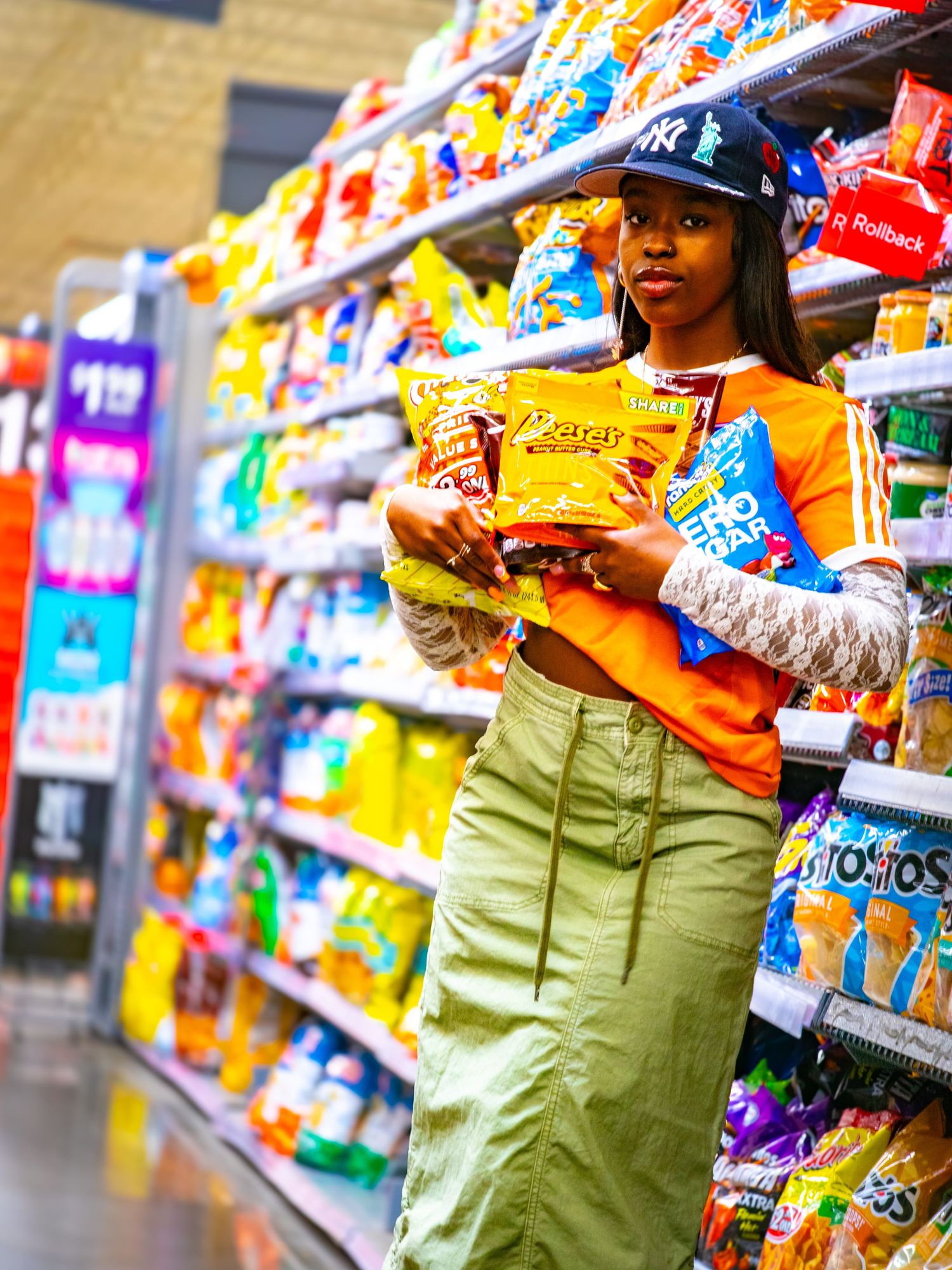 Stylish young woman in orange top and green pants shops for snacks in a colorful grocery store aisle.