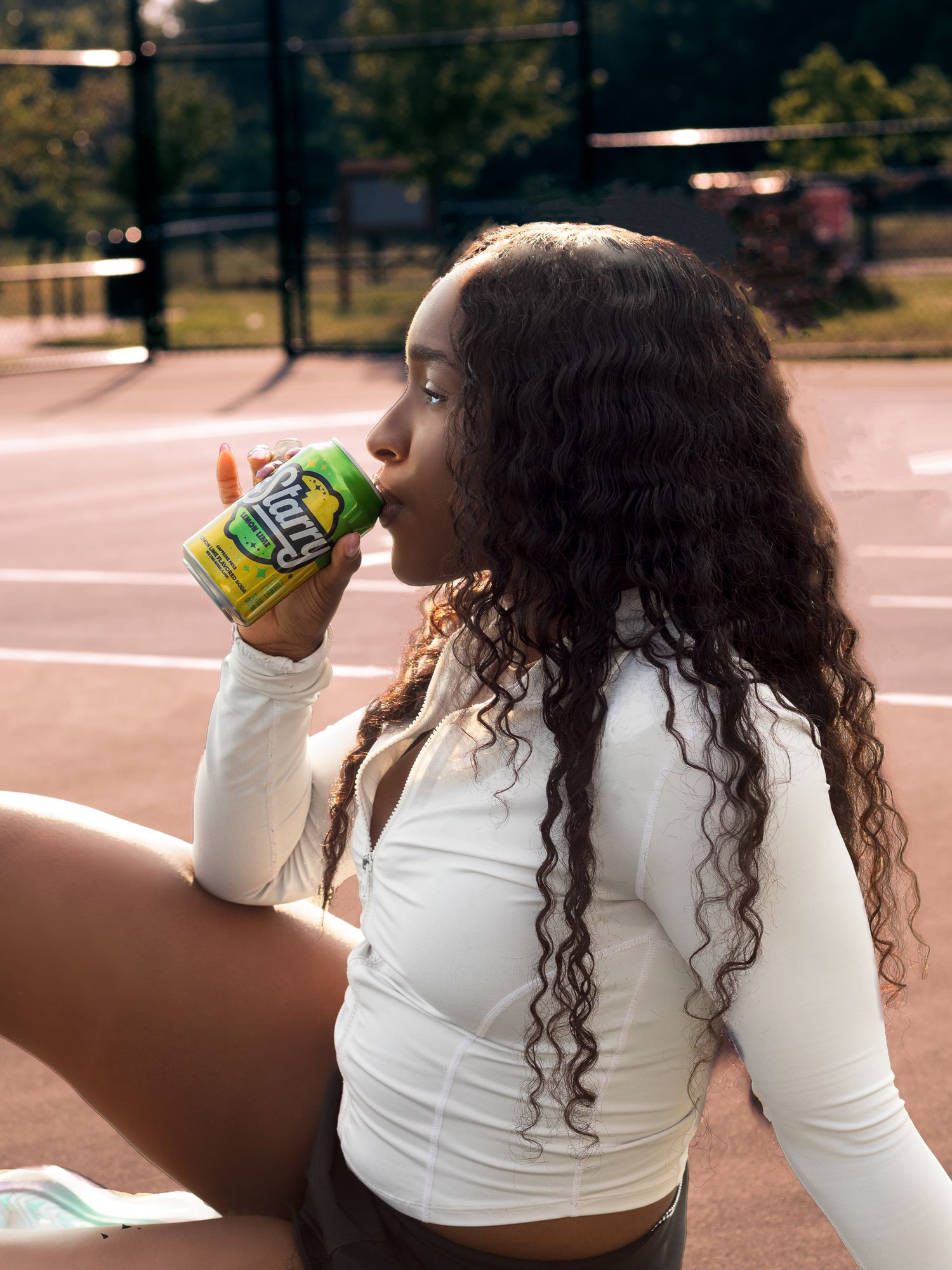 Young woman drinking from a Starry soda can while sitting on an outdoor tennis court in warm light.