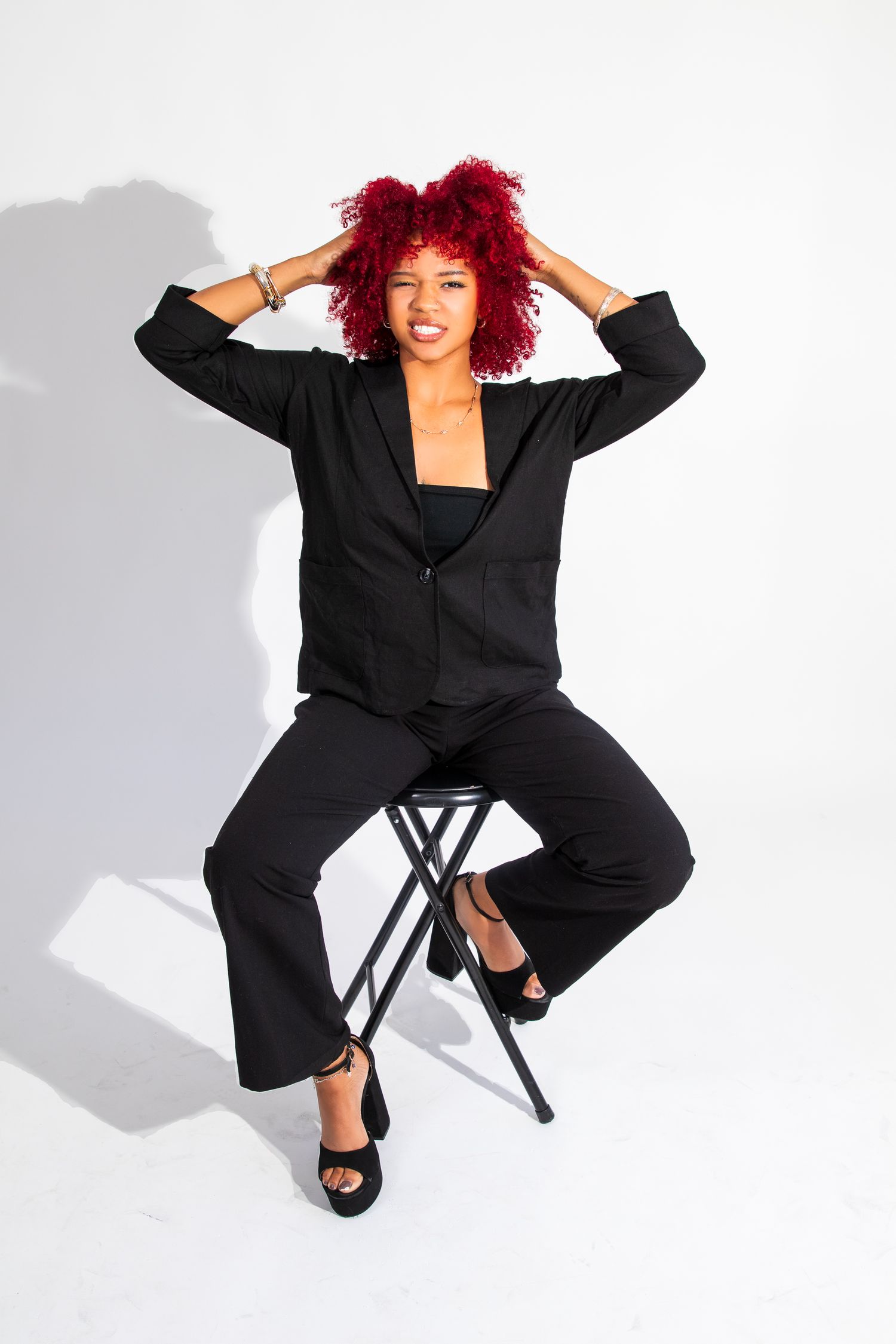 Woman in black suit with vibrant red curly hair poses confidently on stool against white background.