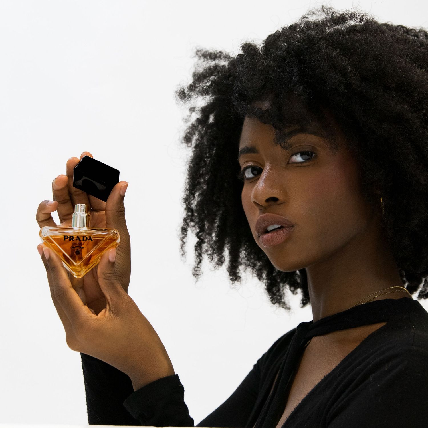 Young Black woman with natural hair holding amber triangle perfume bottle on white background.