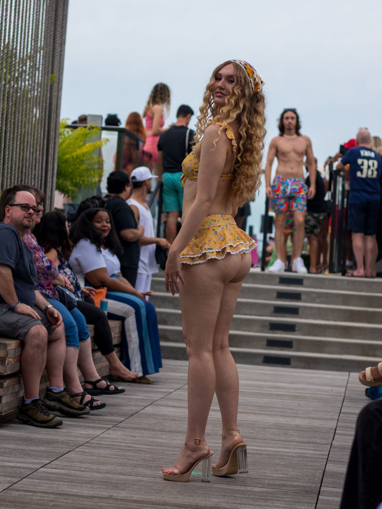 Model in yellow ruffled bikini and headscarf poses showing back view on outdoor fashion runway.