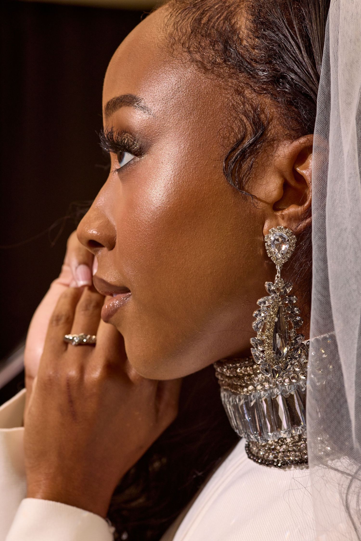 Side profile of Black bride in white veil with large crystal earrings and diamond ring in dark setting.